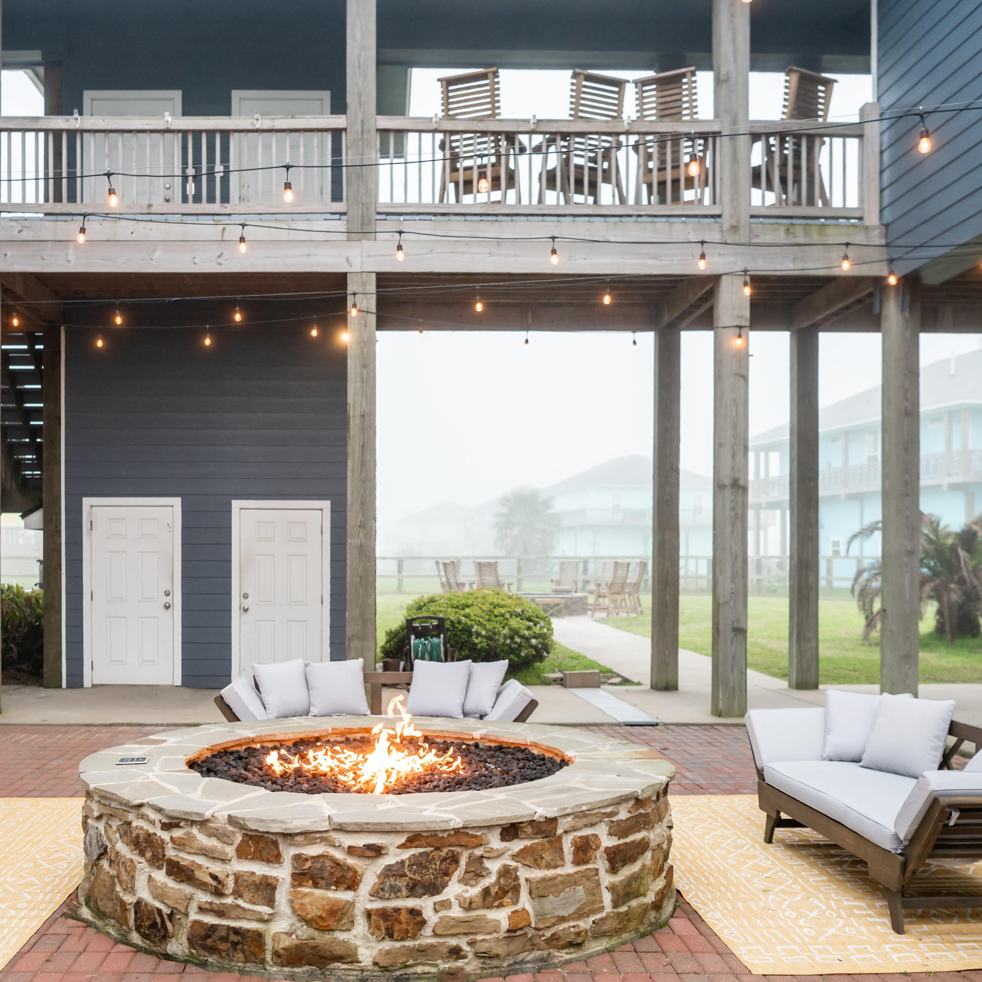 A fire pit surrounded by four lounge chairs, situated under a multi-level deck with string lights against a foggy backdrop.