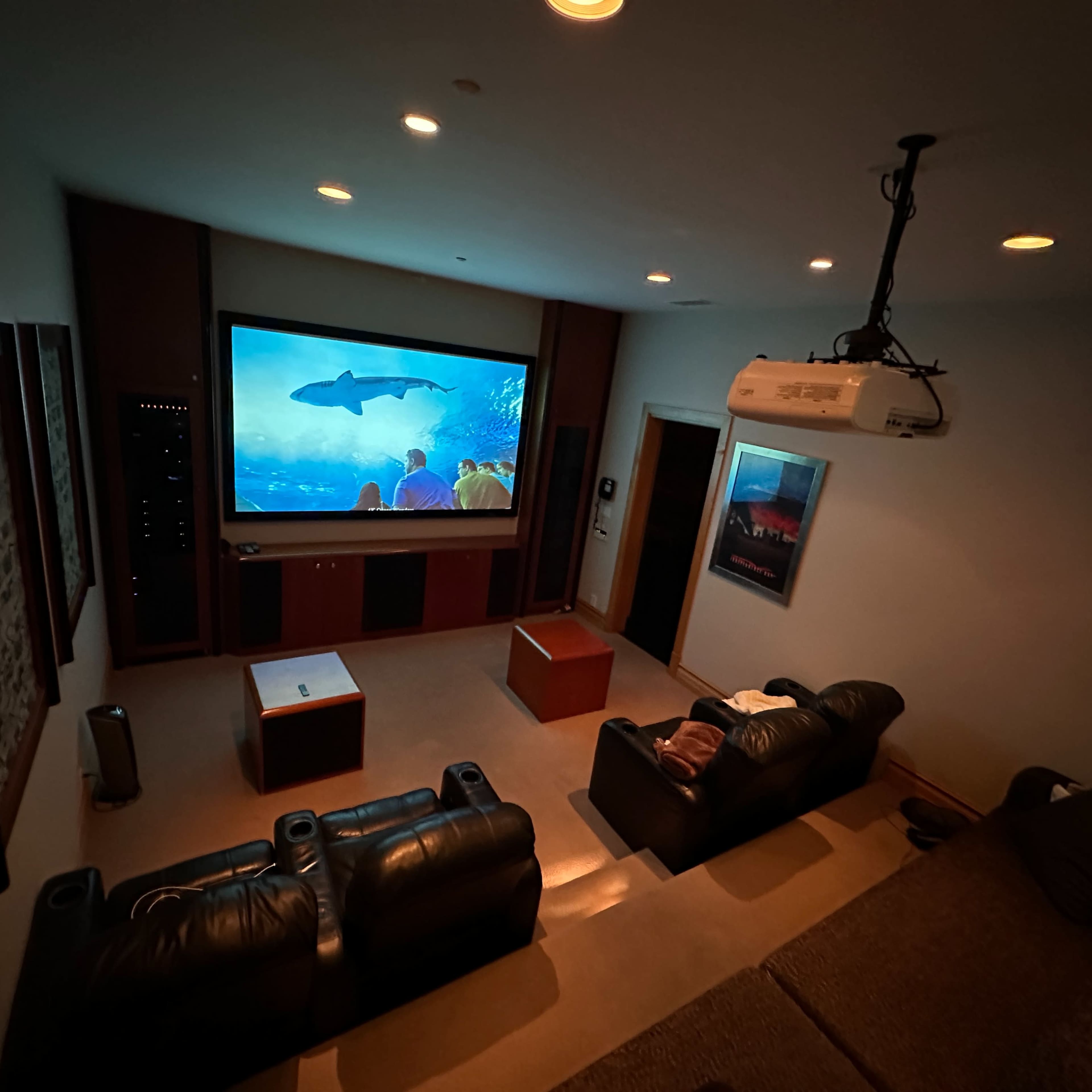 A home theater room features a large projector screen displaying an underwater scene, with black leather seating arranged in front.