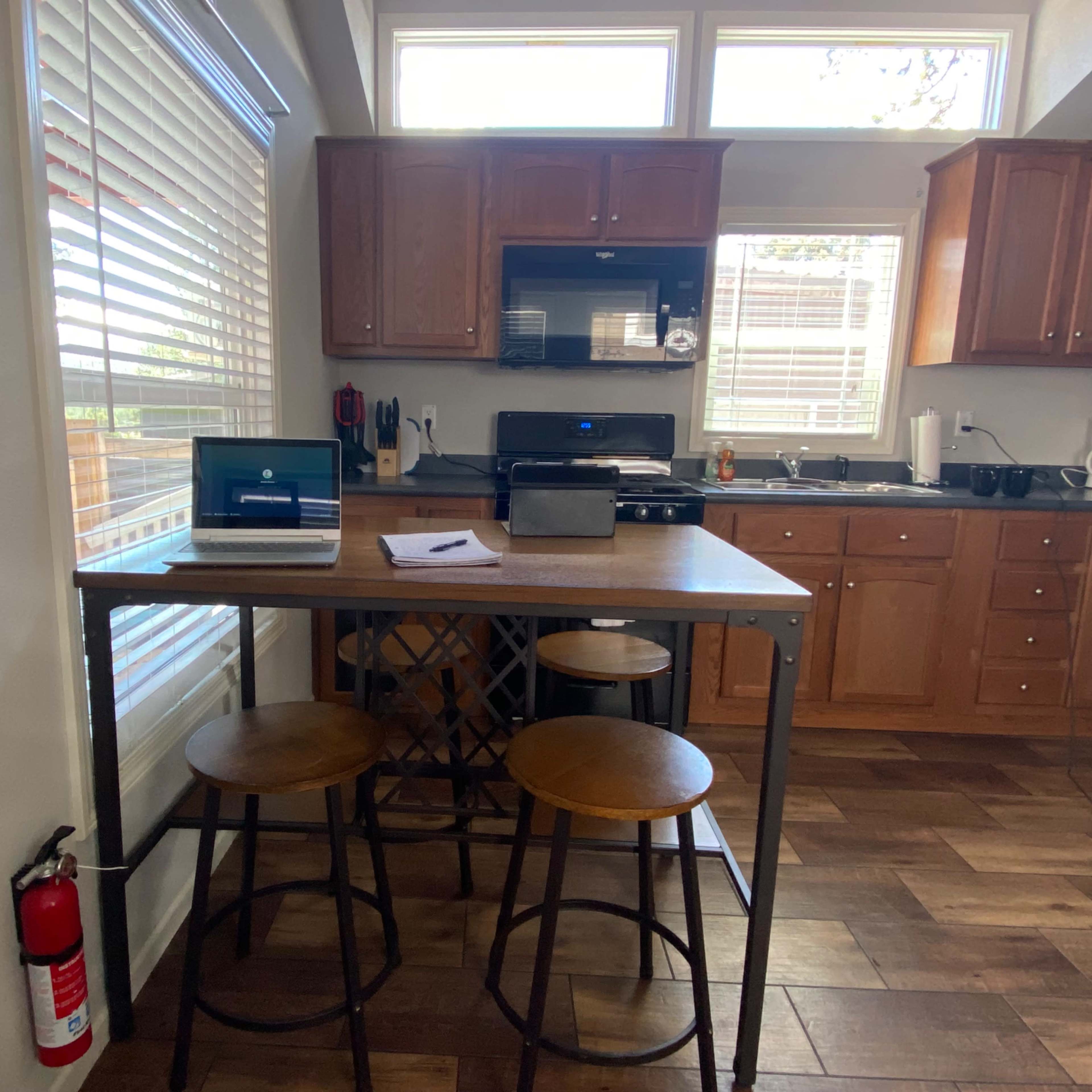 A modern kitchen features wooden cabinets, a countertop with two stools, and large windows allowing natural light.