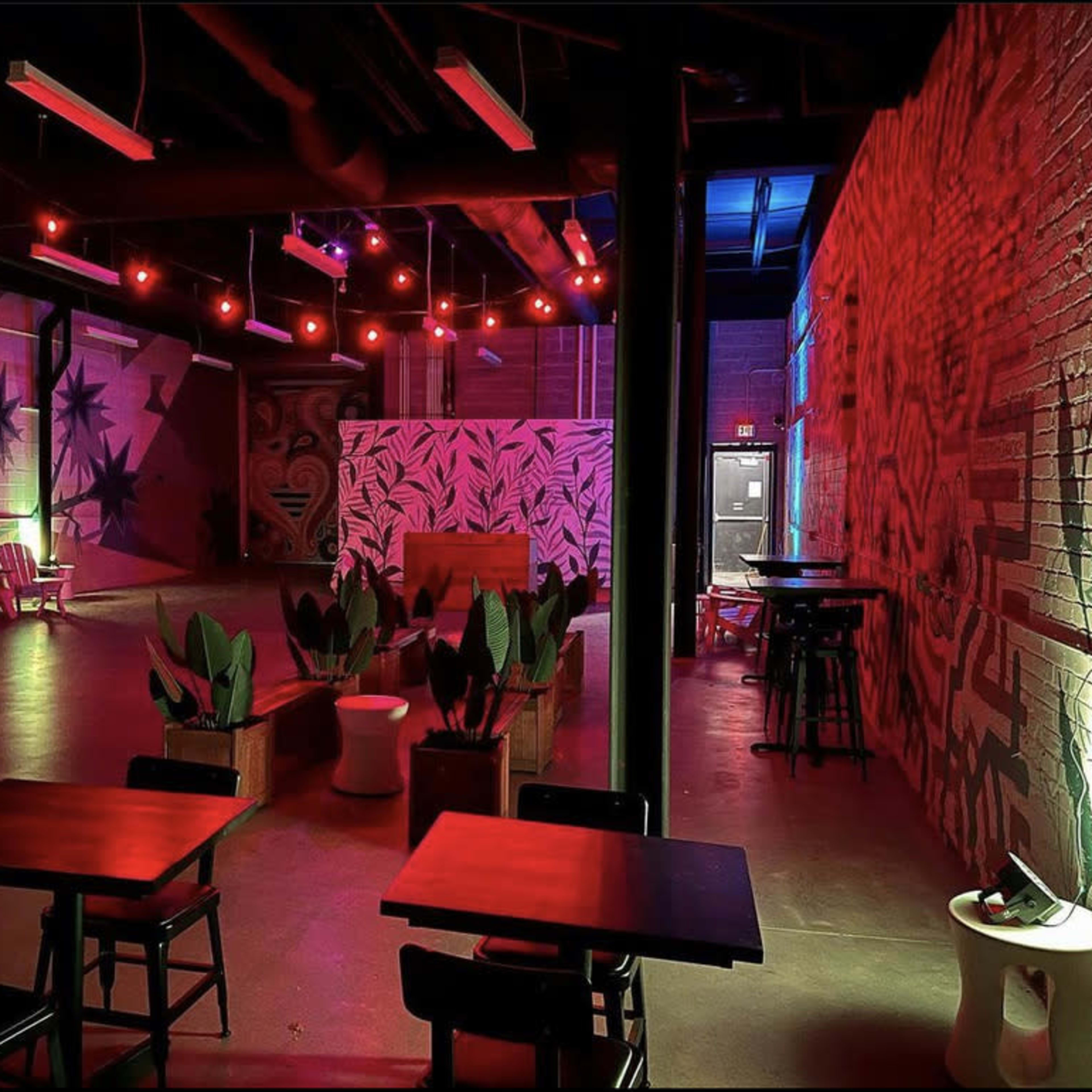 An indoor venue with tables and chairs, decorated walls, and plants, illuminated by colorful lights.
