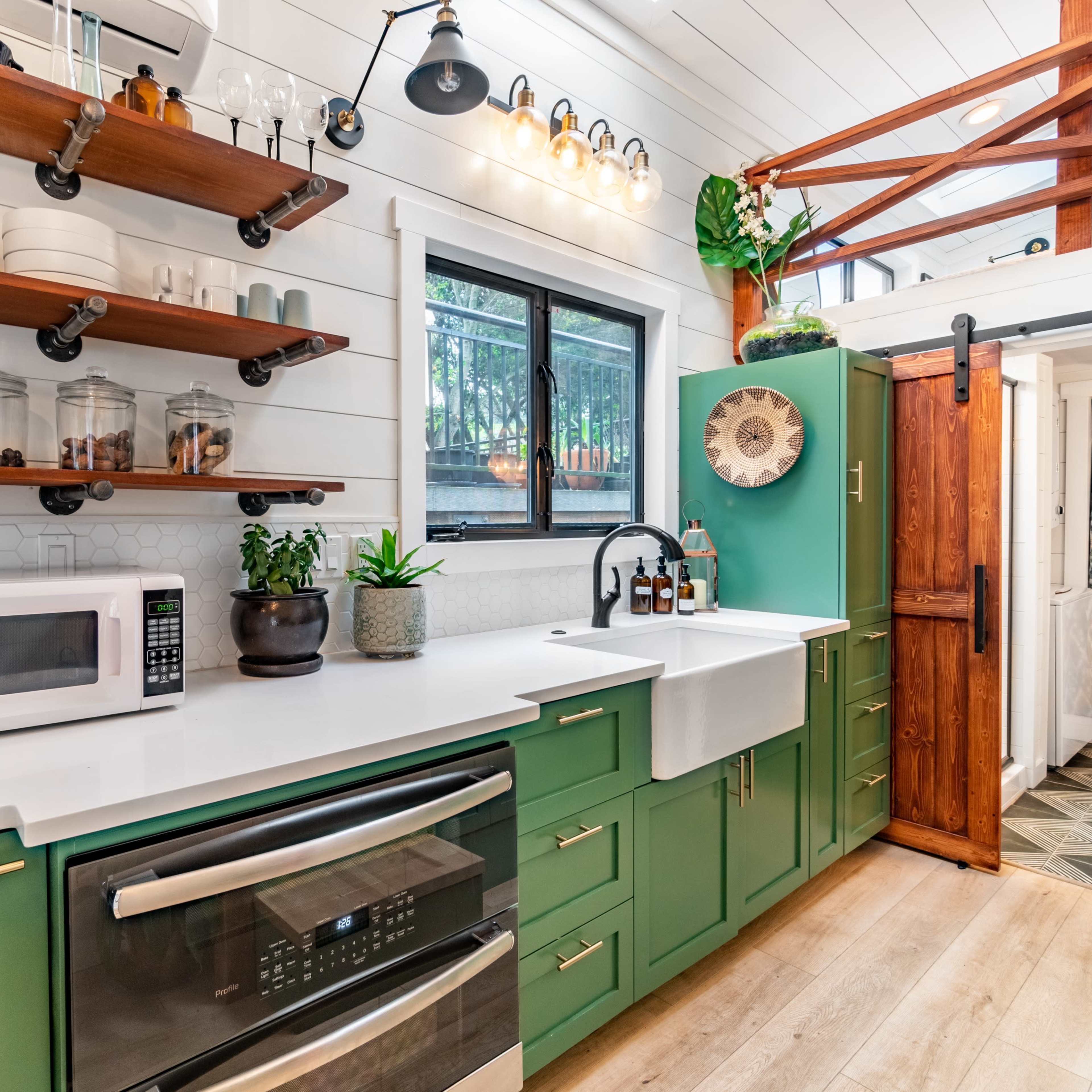 A compact kitchen featuring green cabinetry, open wood shelves with dishware, a farmhouse sink, and a window overlooking a garden.