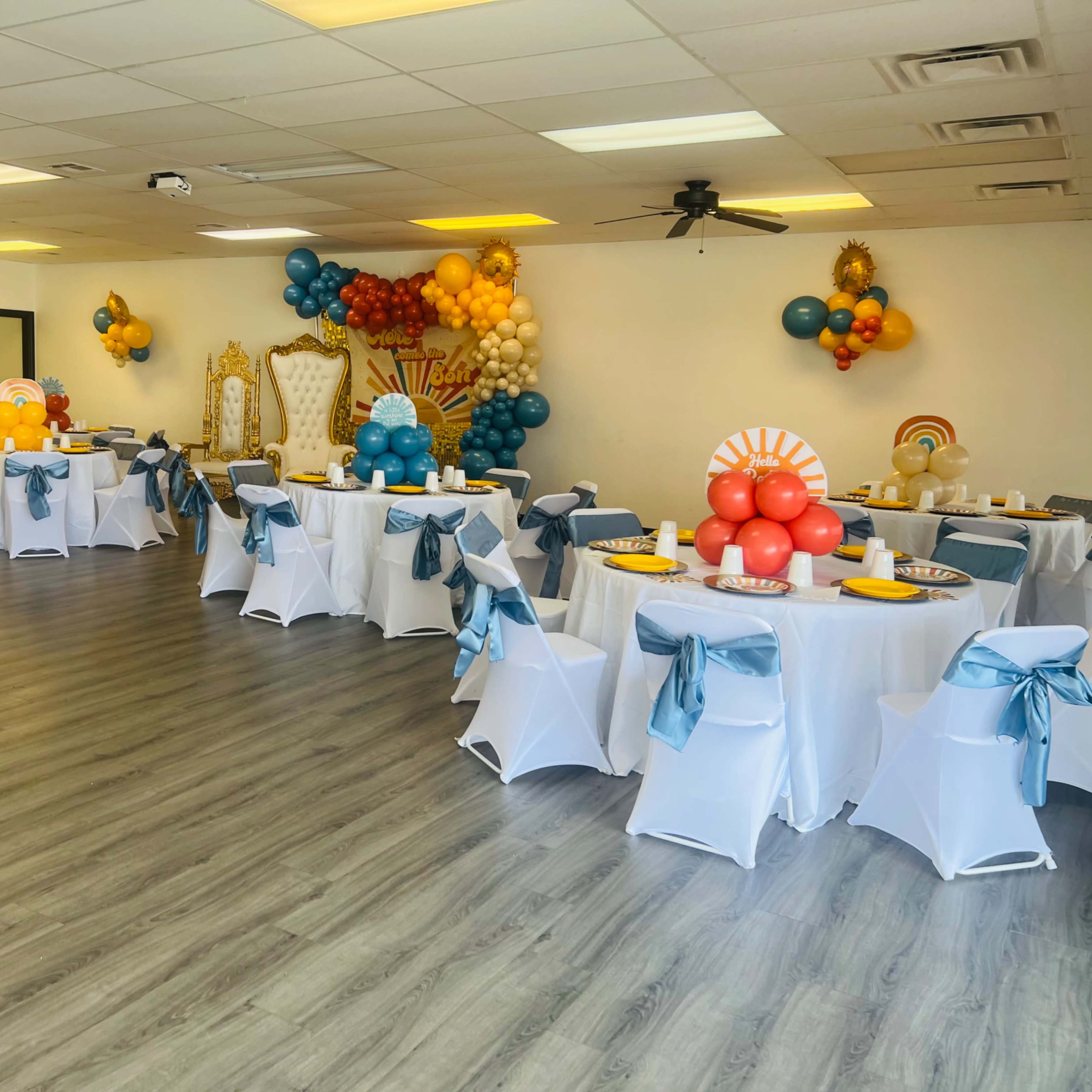 A decorated event space with six circular tables, each covered in white tablecloths and adorned with blue sashes, surrounded by colorful balloon arrangements and a backdrop.