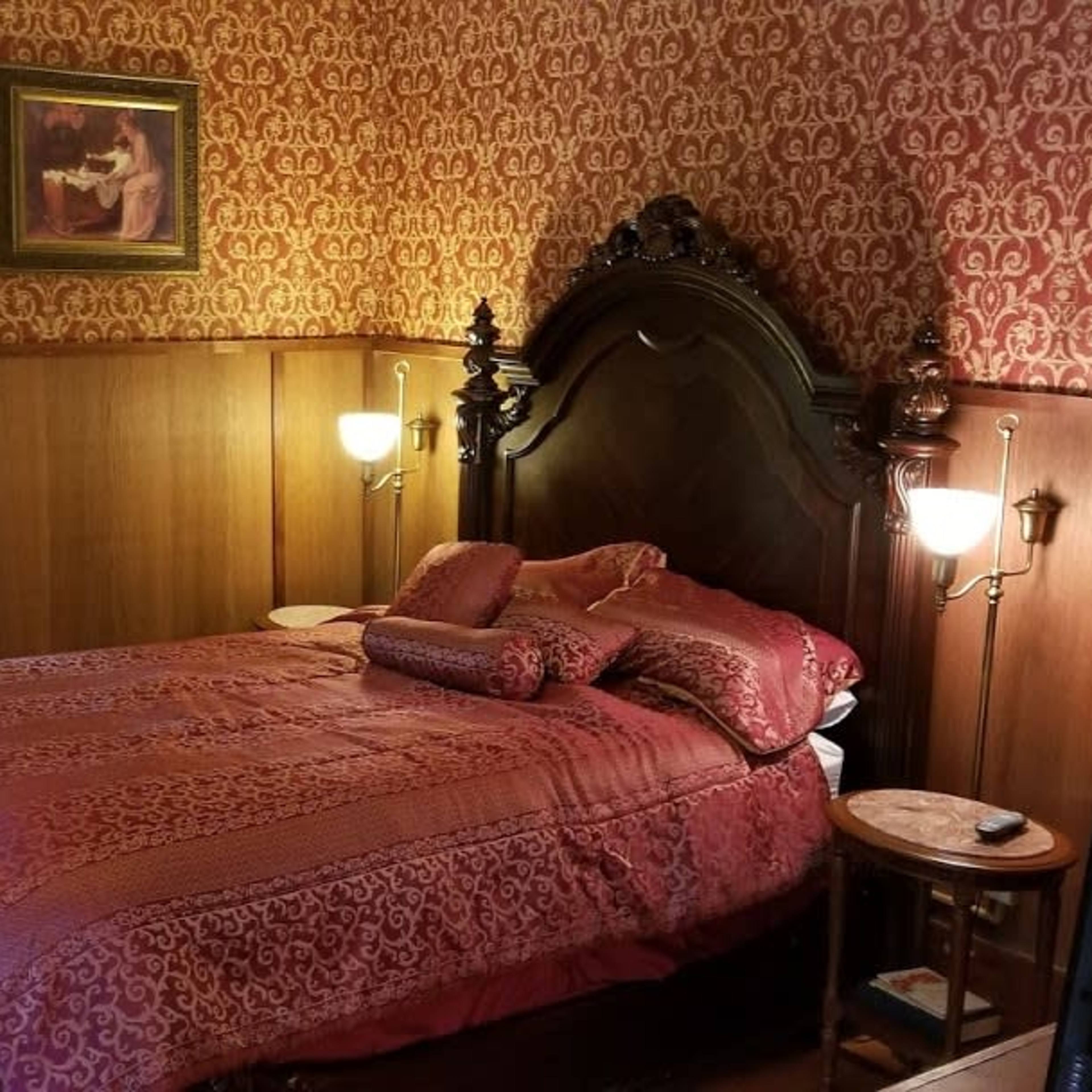 A bedroom with a large bed adorned with red bedding, wooden paneling on the walls, and a window partially covered by a sheer curtain.
