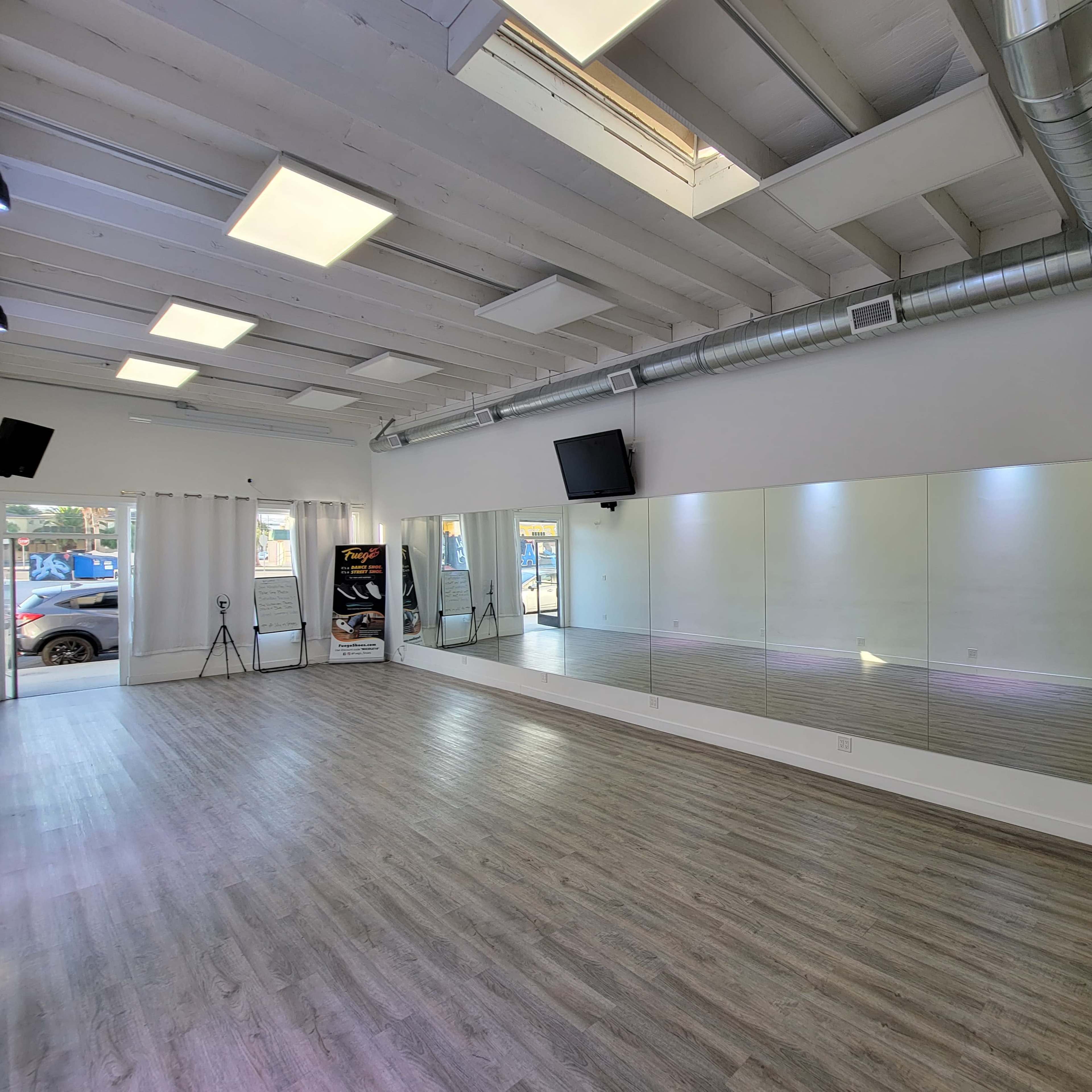 A spacious studio with wooden flooring, large mirrors on one wall, and two wall-mounted speakers.