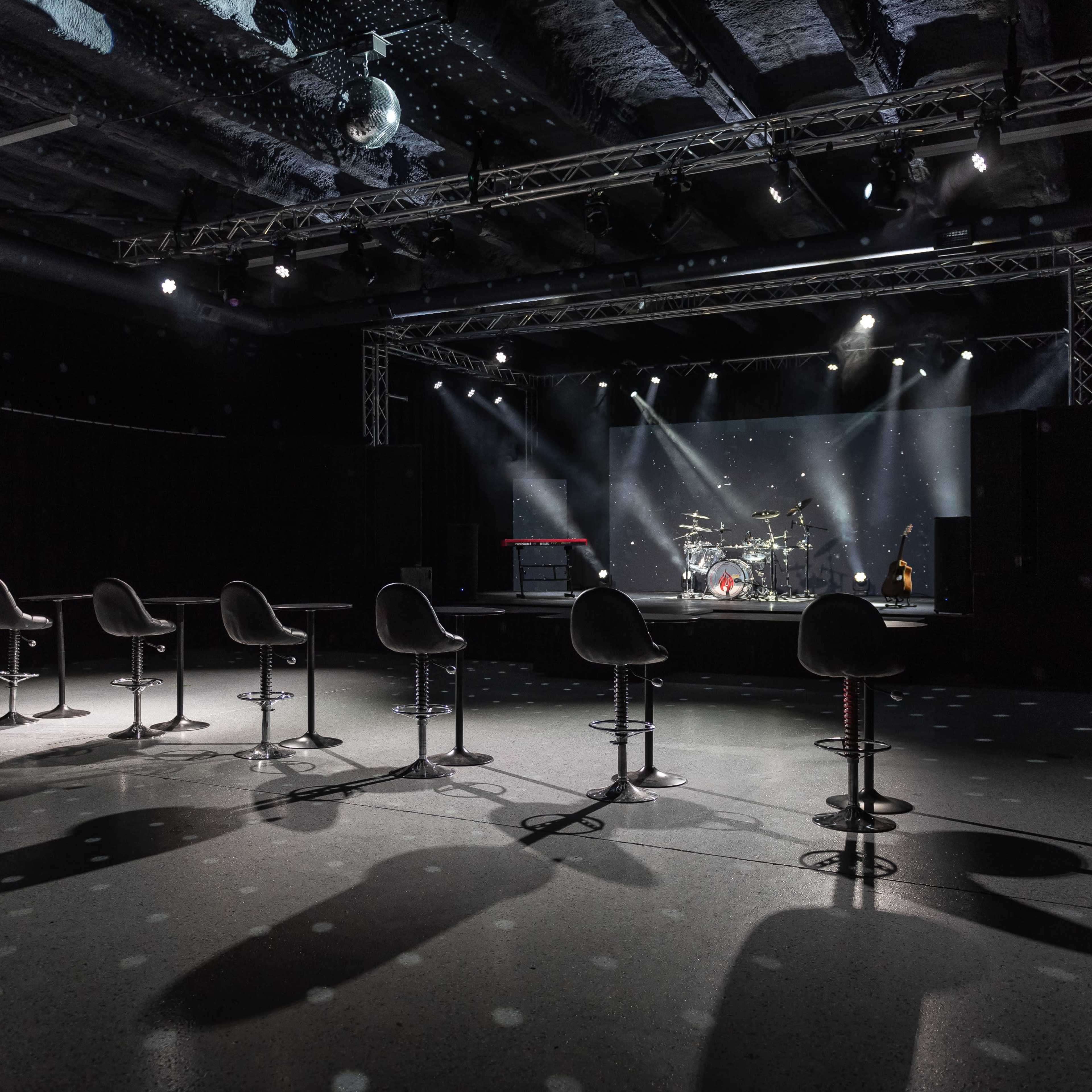 A stage is set up in a dimly lit venue with drum equipment and seven bar stools arranged in front of it.
