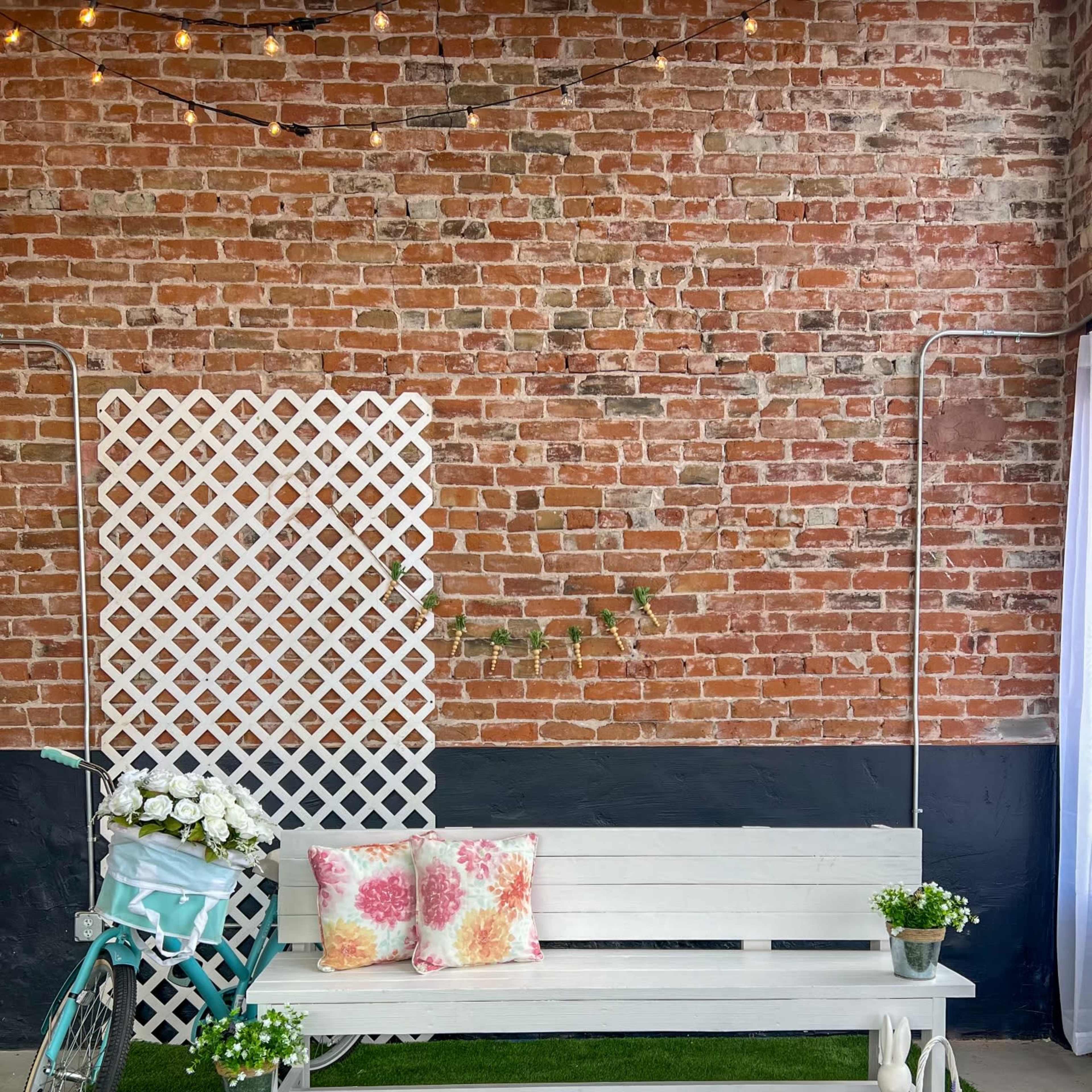 The scene features a white bench with colorful pillows, a bicycle with flowers, and potted plants, set against a brick wall and artificial grass.