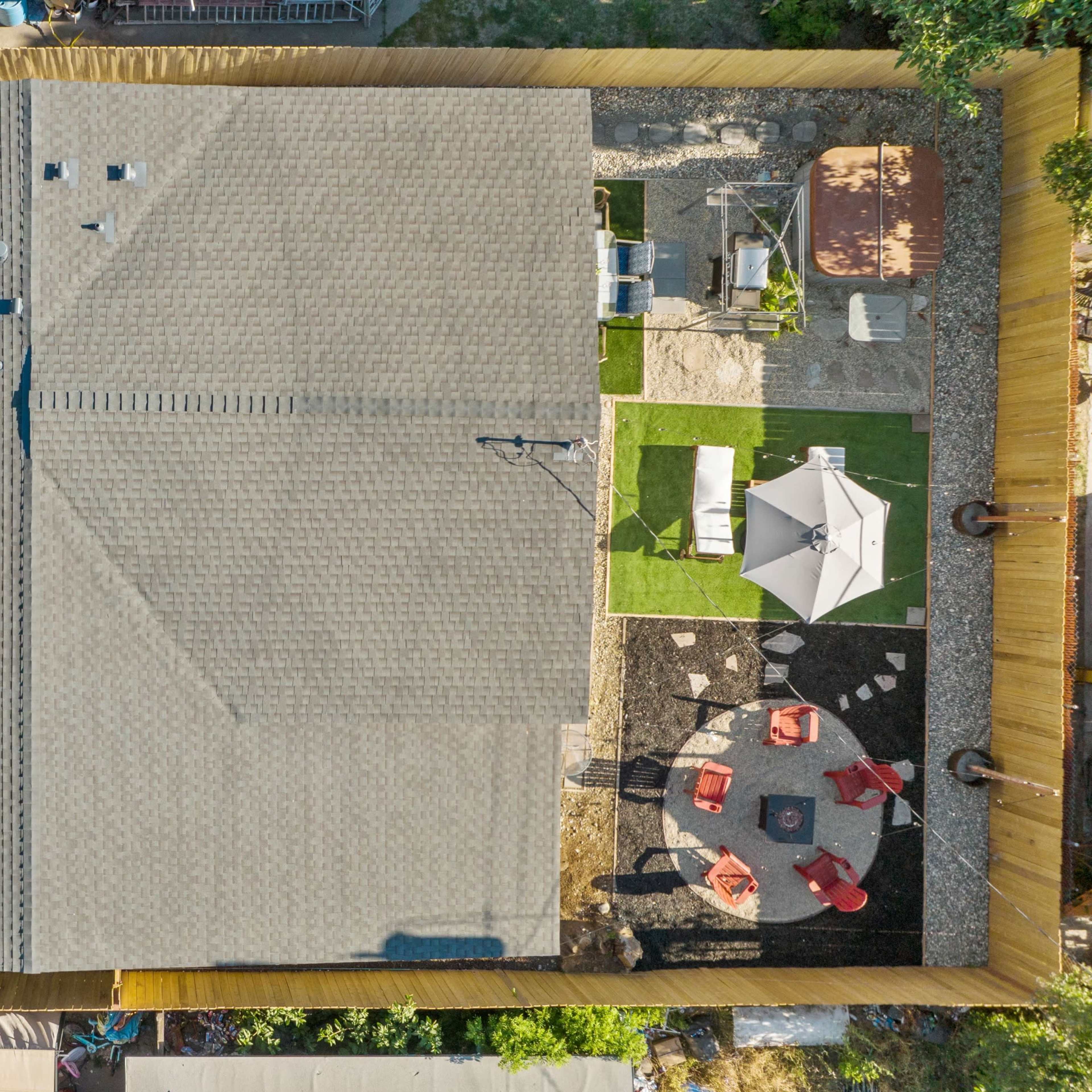 A top-down view of a residential backyard featuring a house with a gray roof, a patio area with red chairs and a table, and green grass surrounded by landscaped sections.