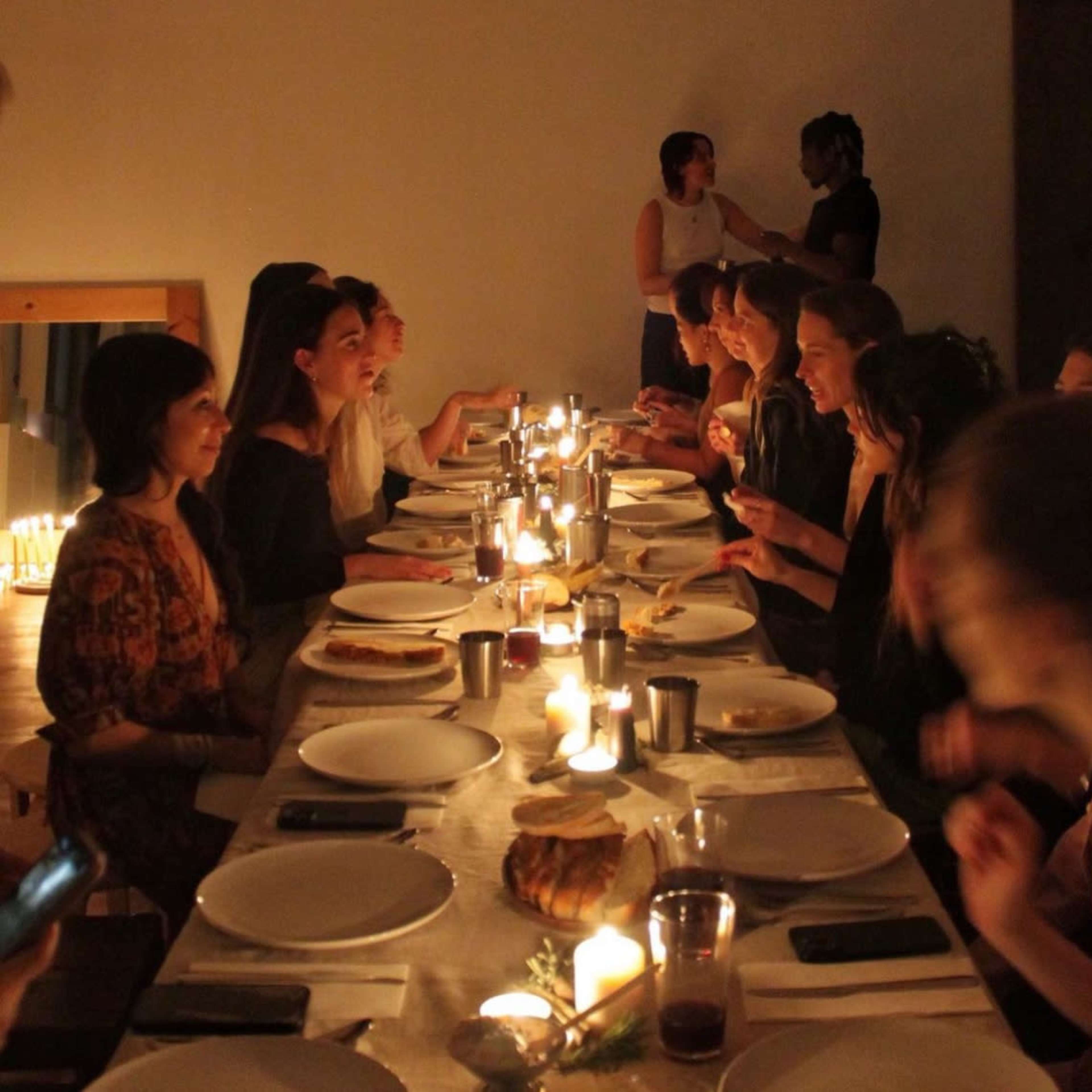 A long dining table is set with plates and candles, with a group of people engaged in conversation during a dinner gathering.