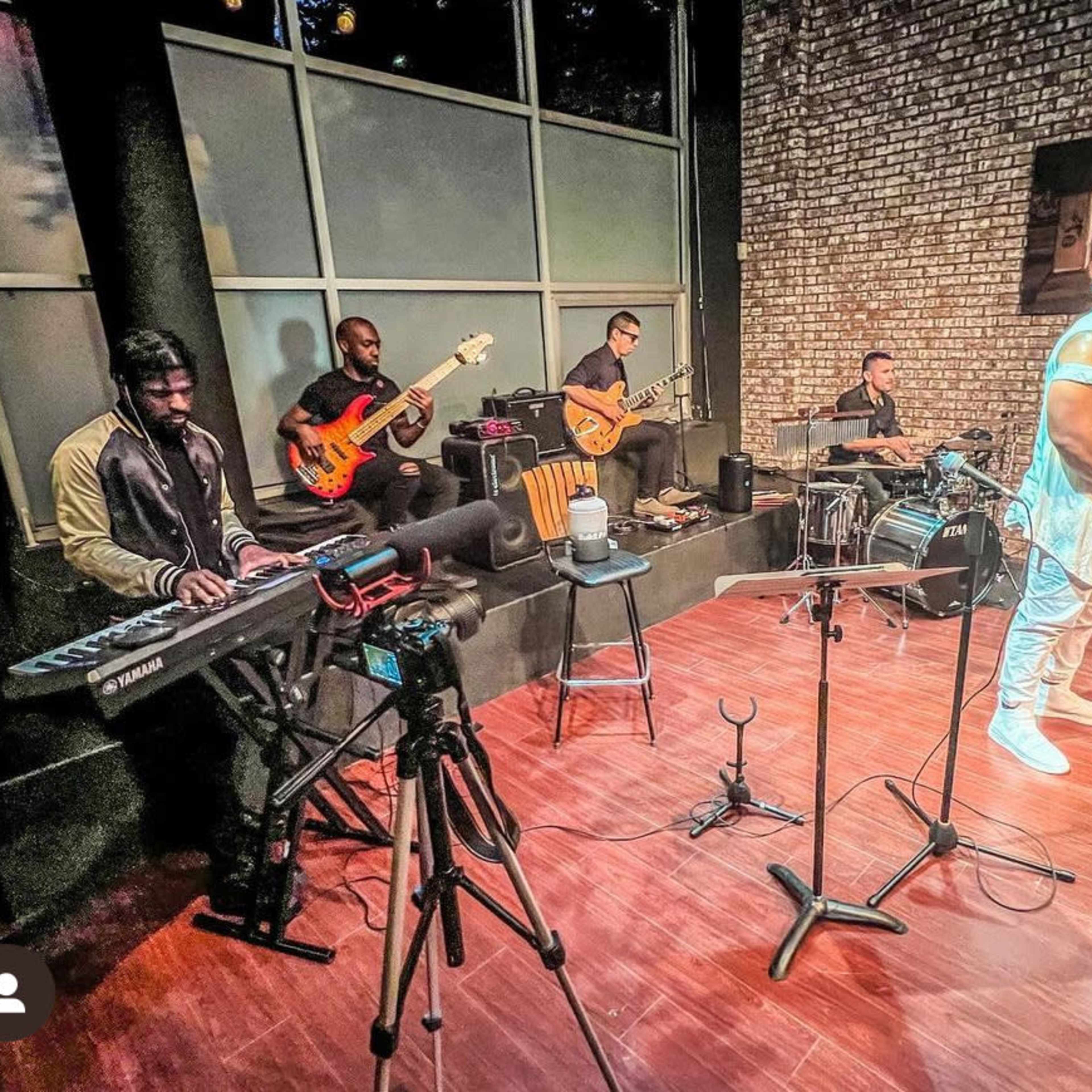 A group of musicians performs on a stage featuring a saxophonist, a keyboardist, a drummer, and two guitarists in a brick-walled setting.