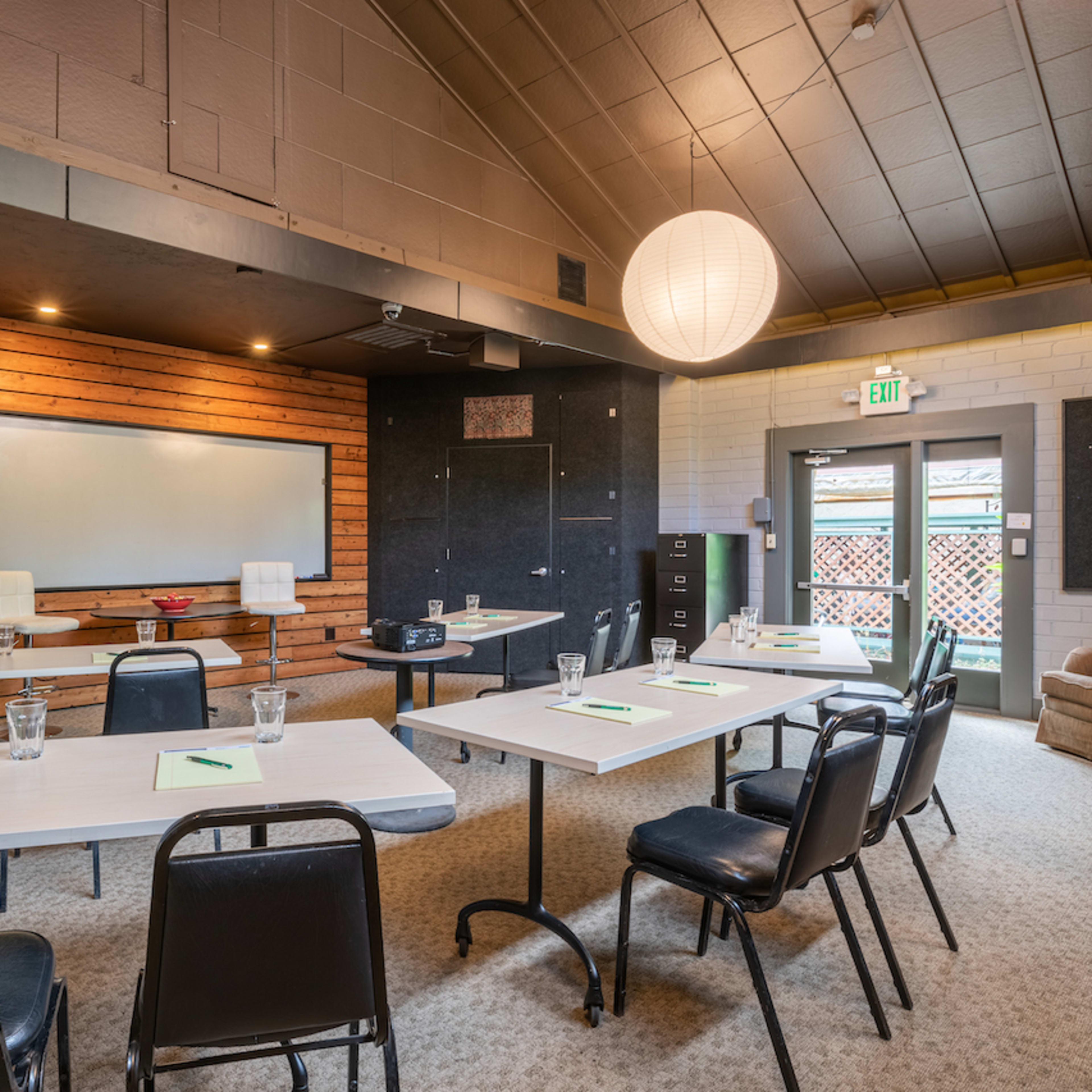 A meeting room with several tables and chairs arranged for a presentation, featuring a whiteboard and a large pendant light overhead.