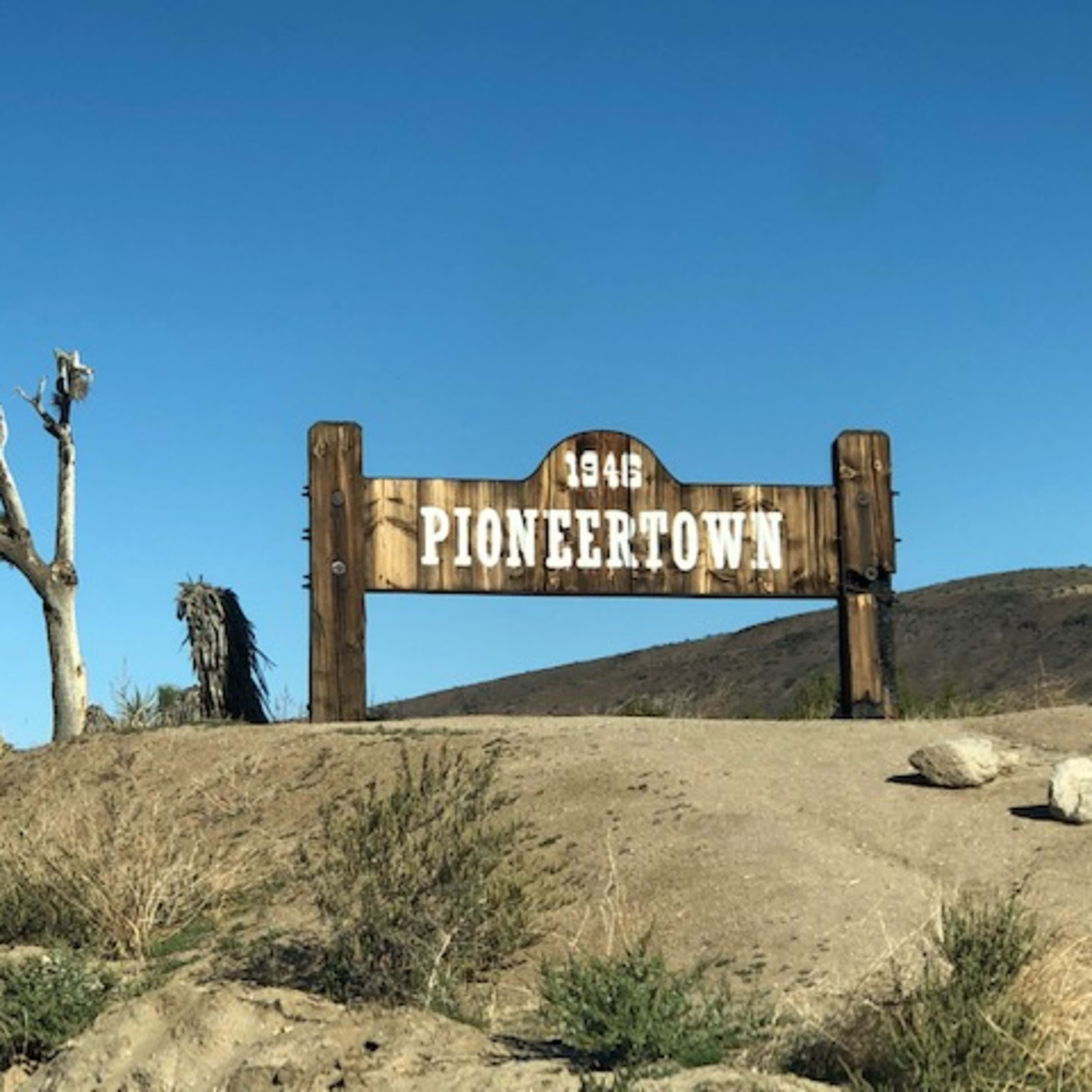 A wooden sign reads "Pioneertown 1946" beside a sparse, dry landscape and a lone tree.