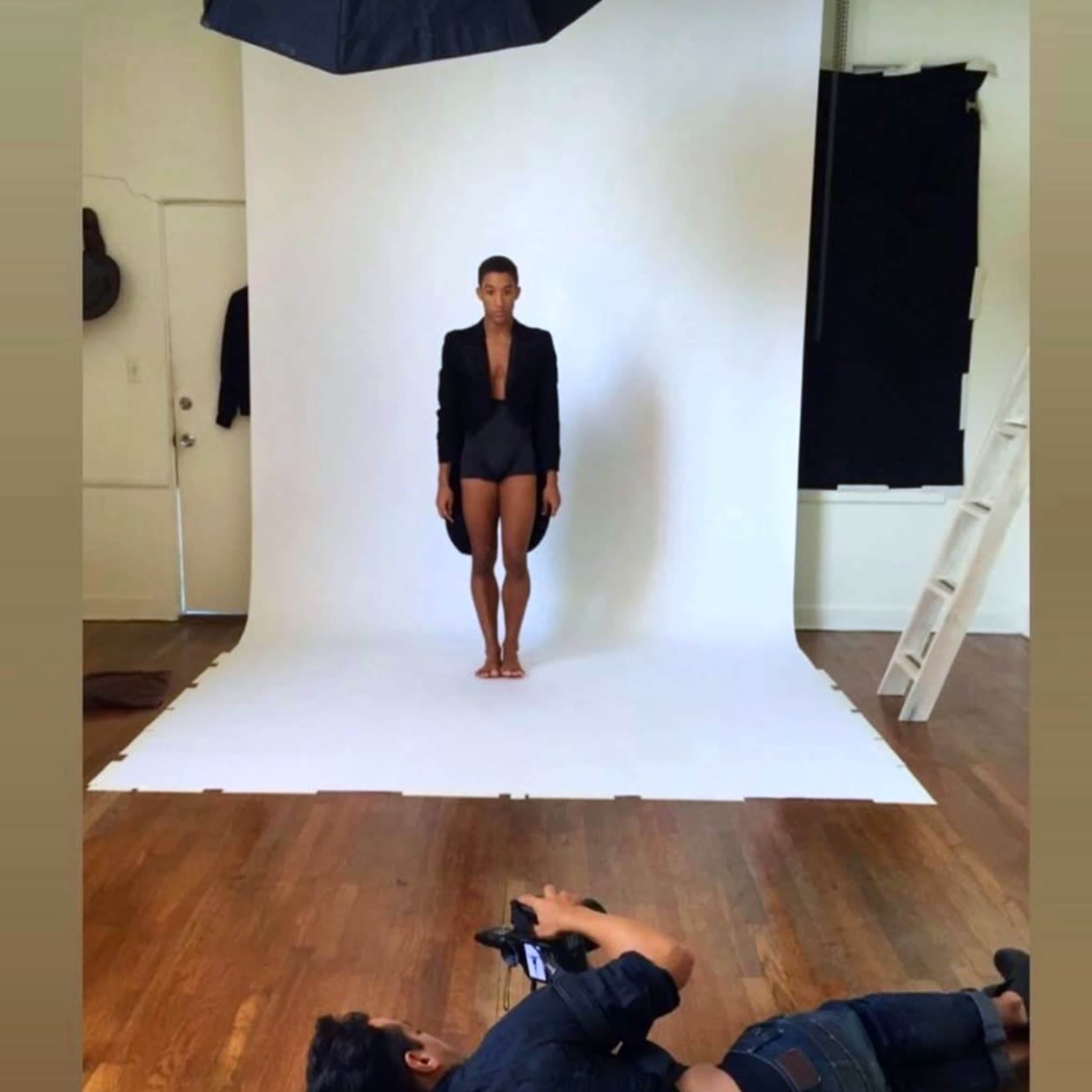 A model stands on a white backdrop in a photography studio while a photographer captures images from a kneeling position.