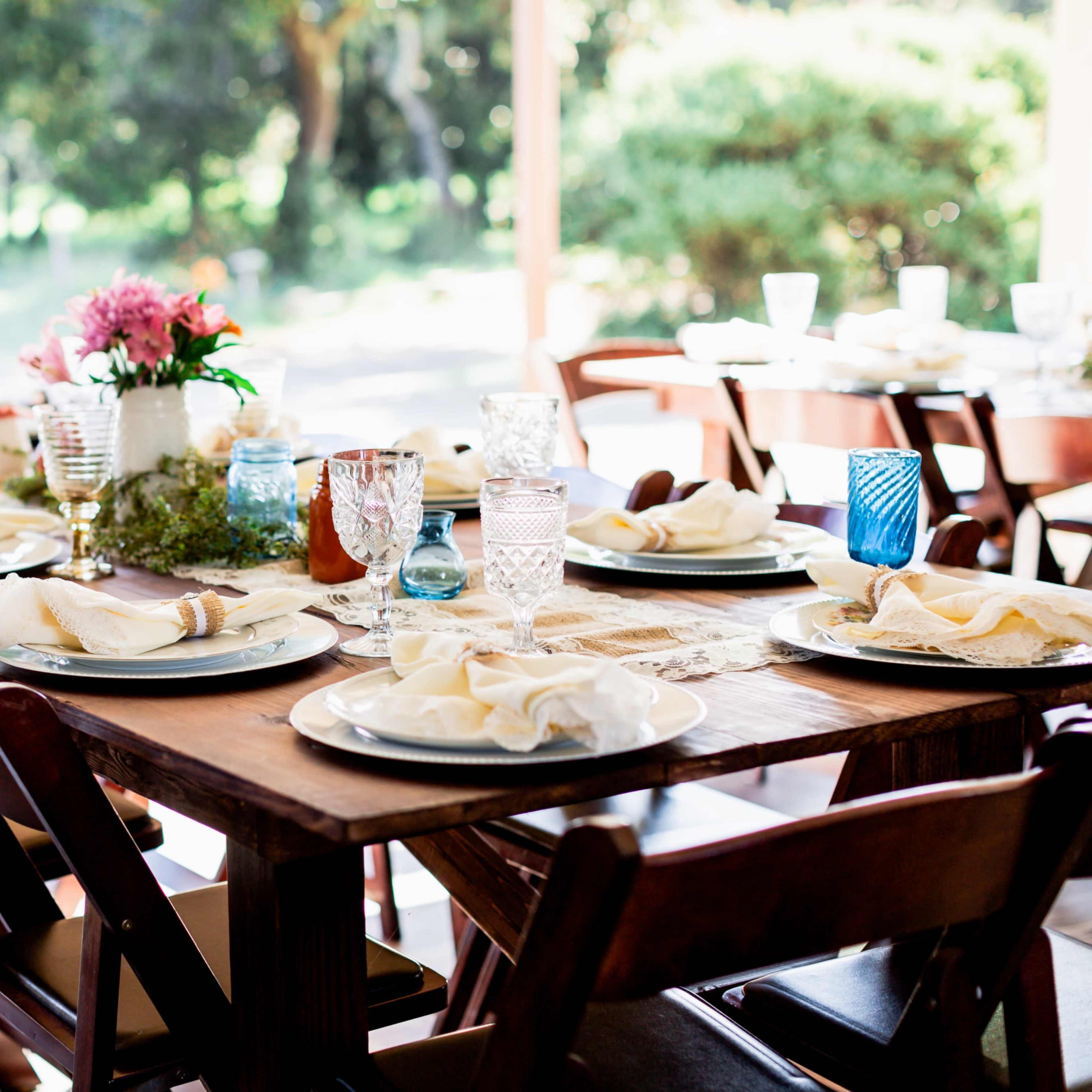 A long wooden dining table is set with plates, napkins, and decorative glassware, surrounded by chairs, in a bright outdoor setting.