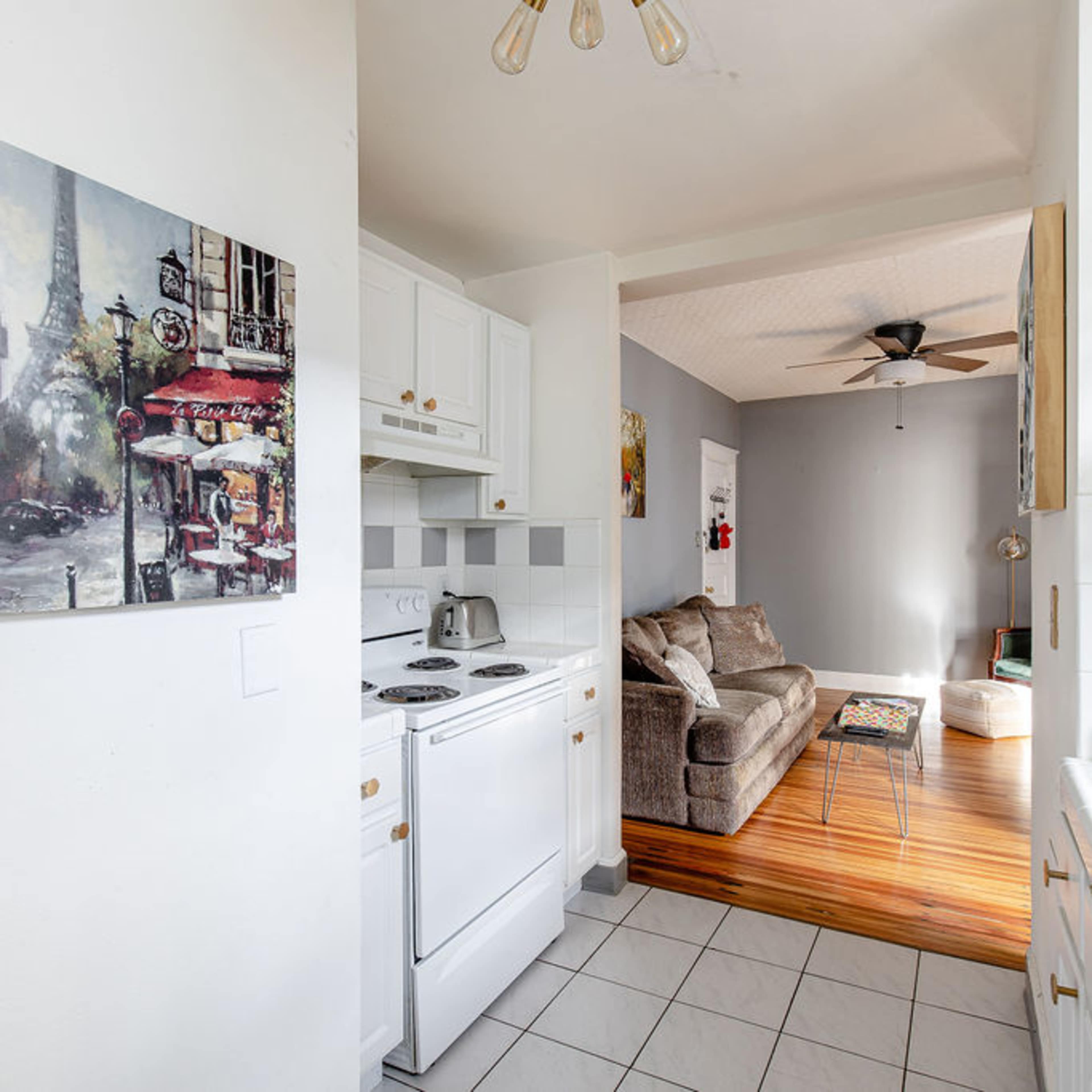 A kitchen area with a white stove and cabinets leading into a living room featuring a gray wall and a couch.