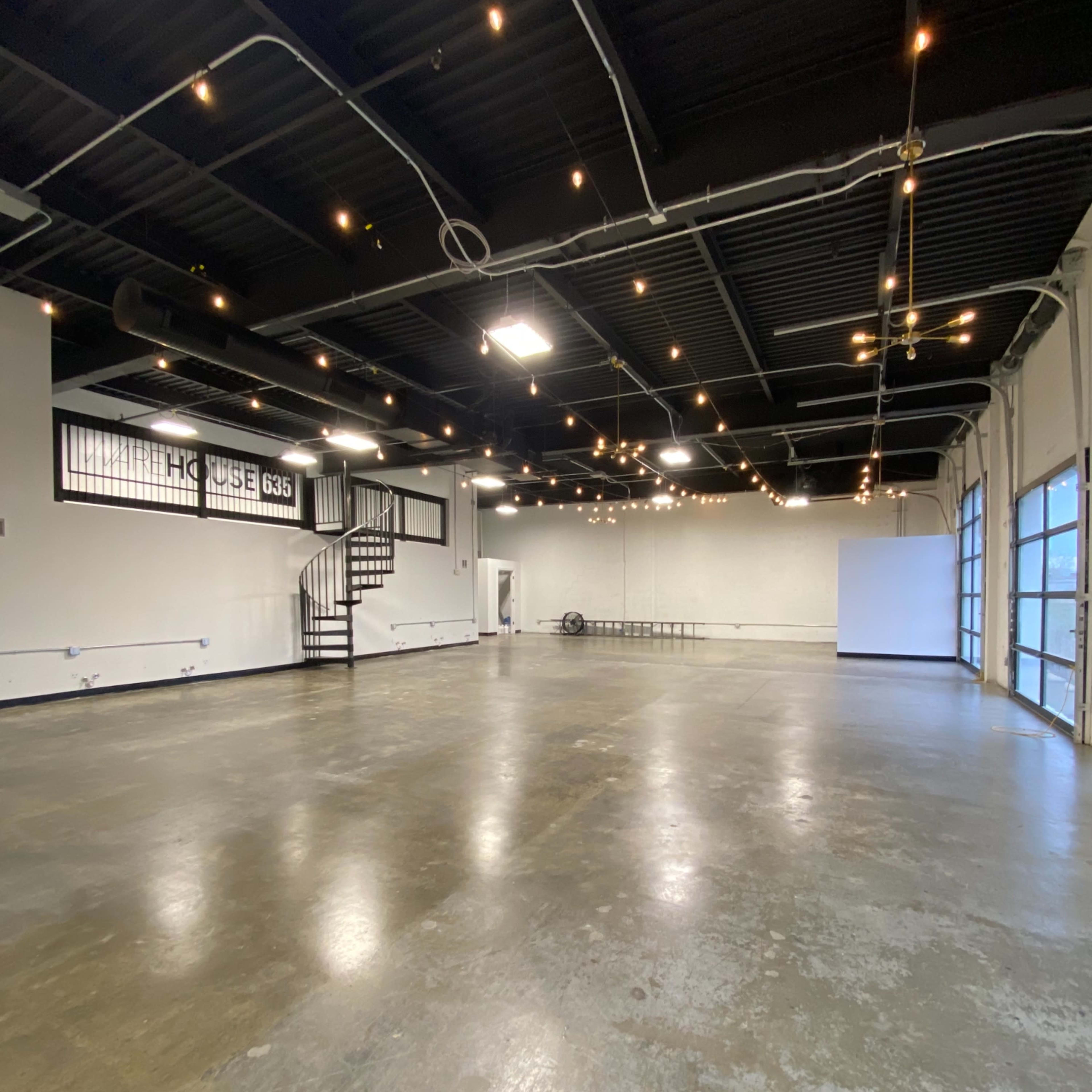 An empty industrial space with concrete flooring, exposed ceiling beams, and string lights overhead.