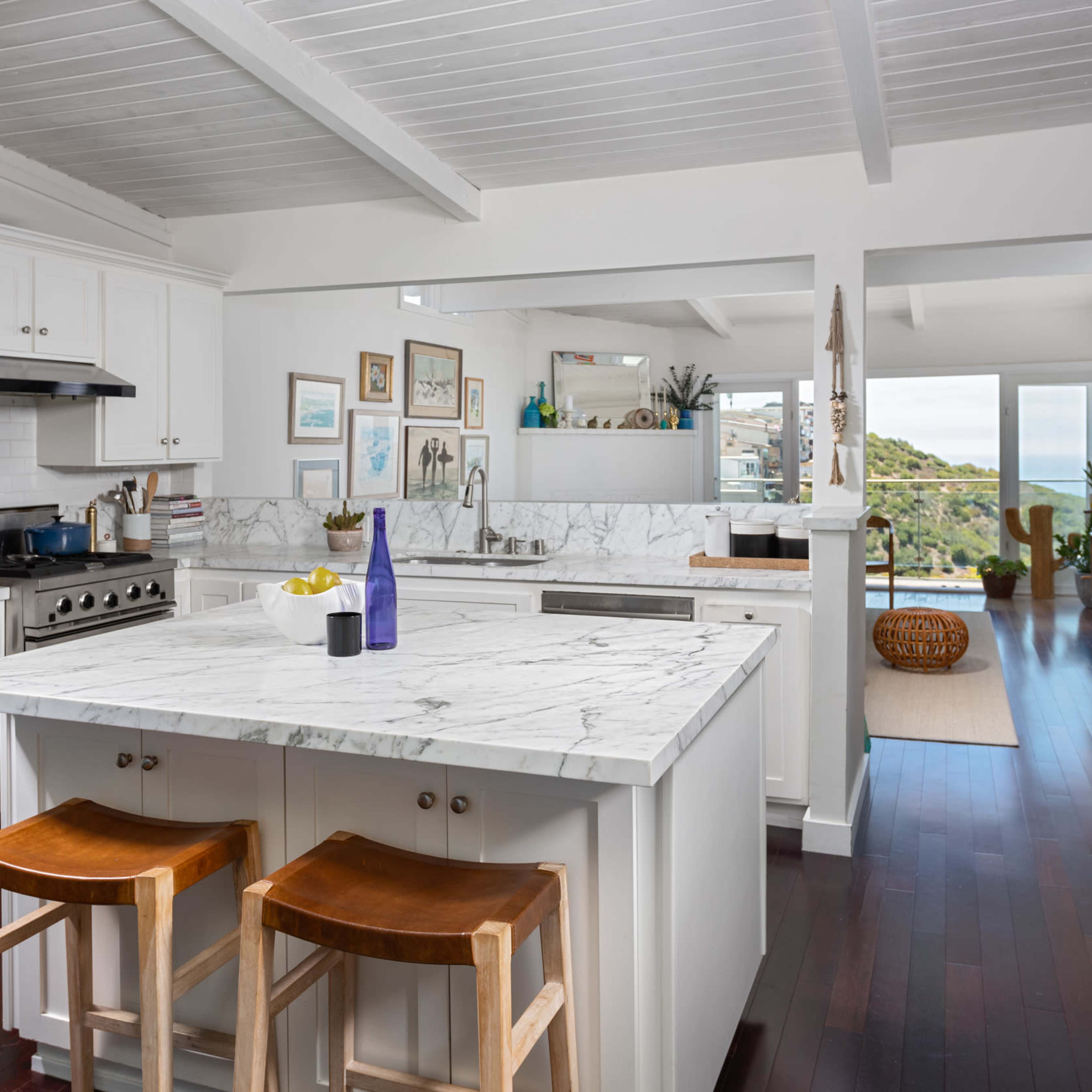 A modern kitchen features white cabinetry, a marble-topped island with two wooden stools, and large windows offering a view of the outdoor scenery.