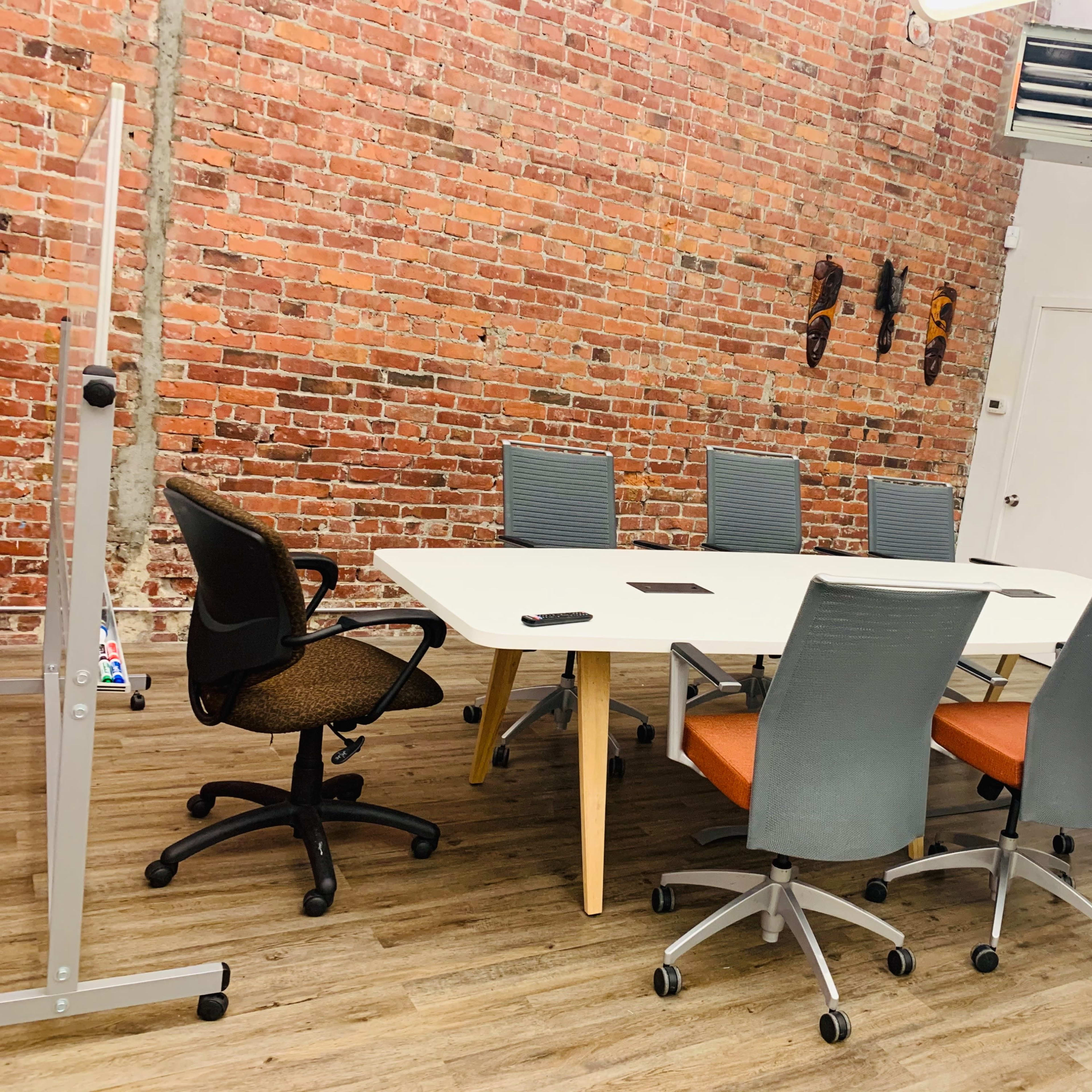 A modern conference room with a long white table surrounded by six office chairs, a brick wall in the background, and a wall-mounted screen.