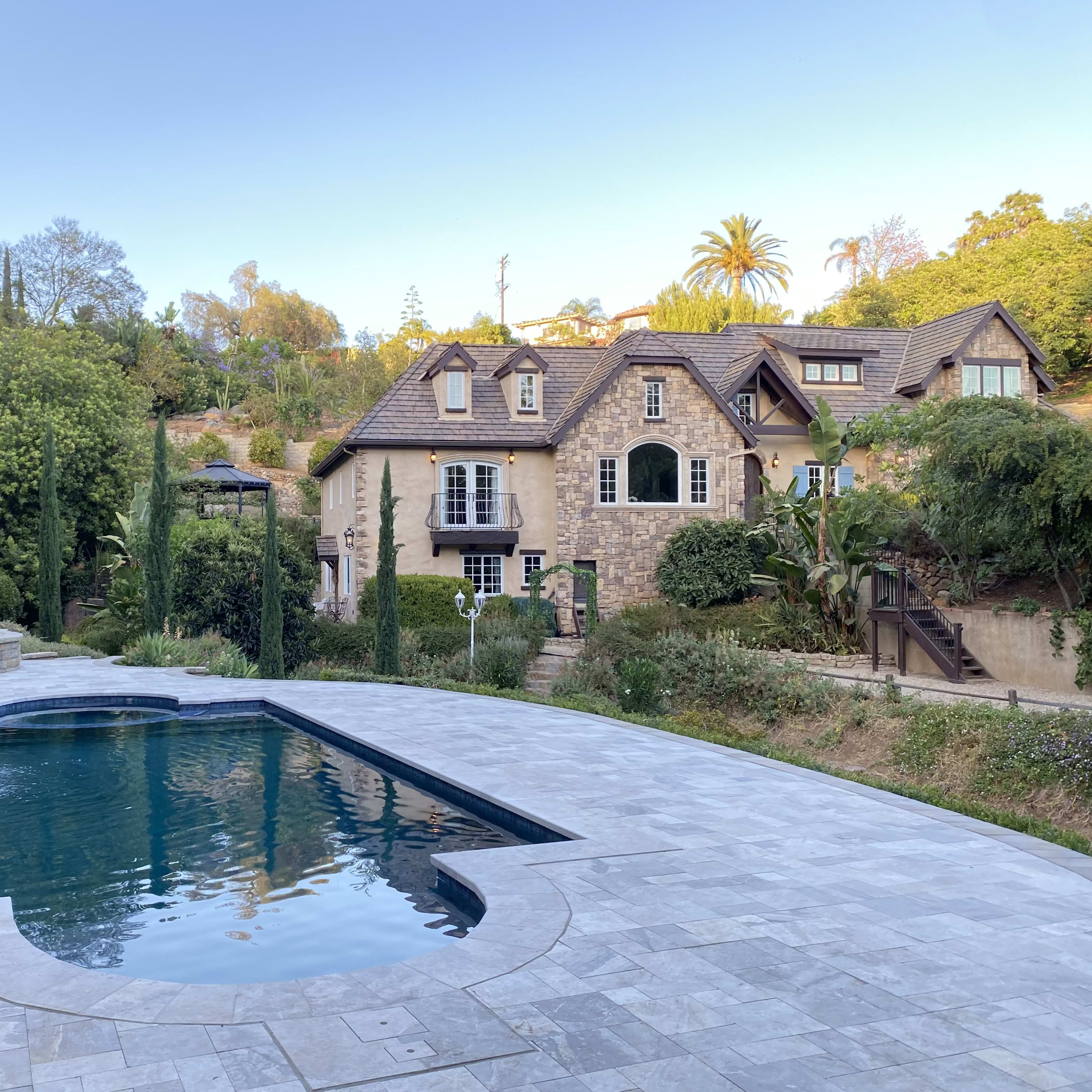 A large stone house next to a rectangular swimming pool, surrounded by trees and landscaping.
