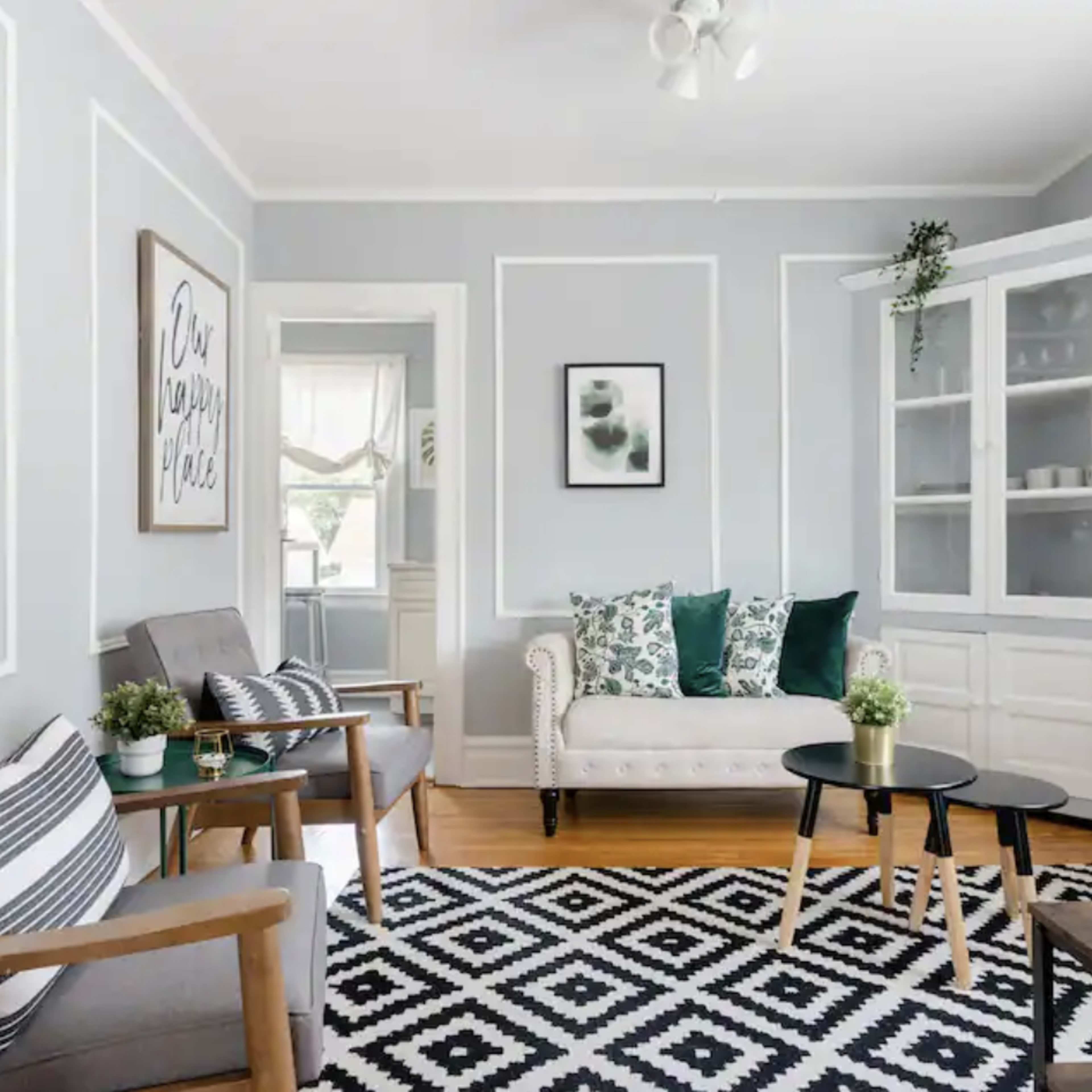 A living room featuring a light gray color scheme, a striped black and white area rug, a white sofa with green cushions, and two wooden chairs beside a small black coffee table.