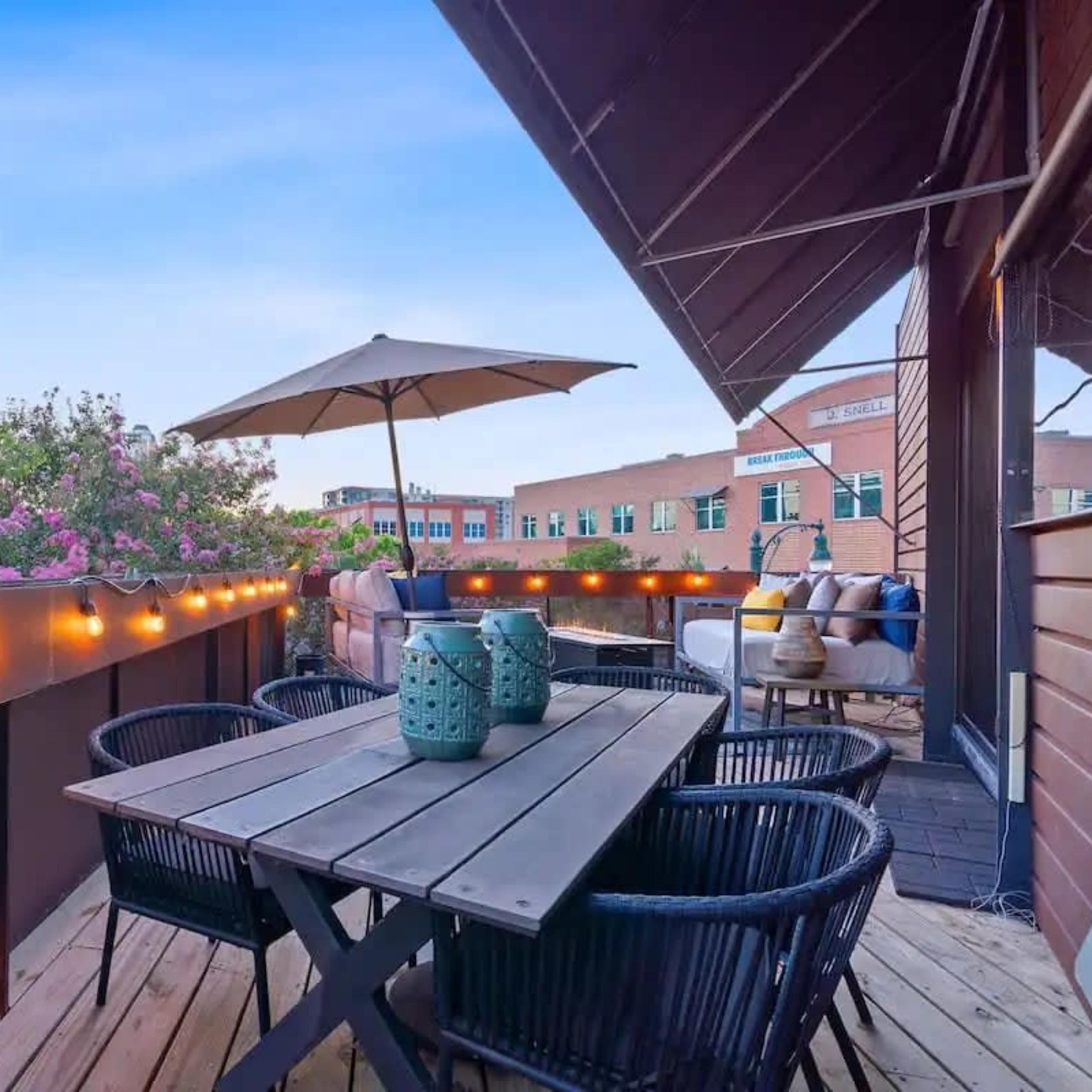 A rooftop terrace features a wooden table surrounded by black wicker chairs, with string lights and an umbrella, overlooking greenery and a brick building.