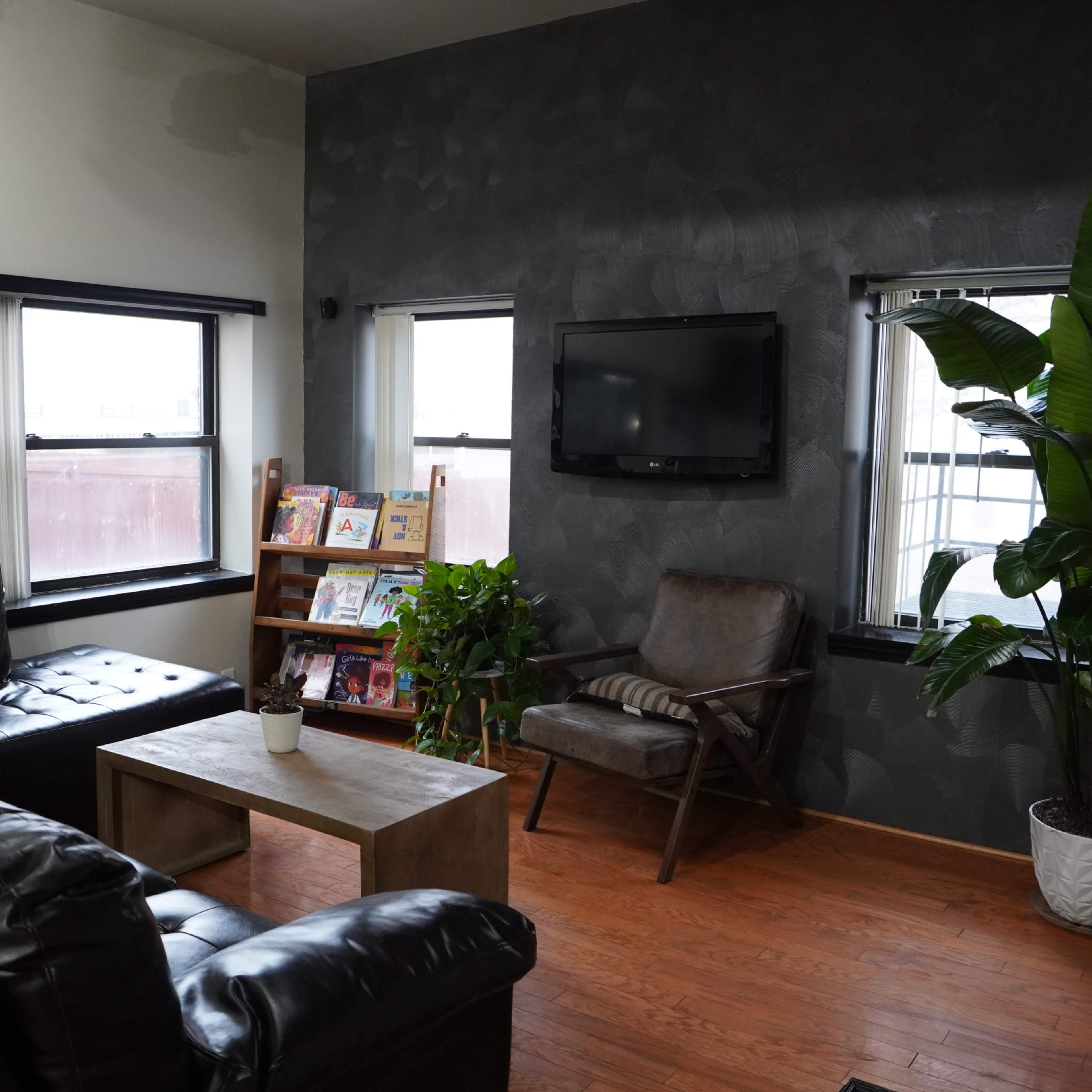 A cozy living room with a black leather couch, a wooden coffee table, a plant next to an armchair, and a wall-mounted television.