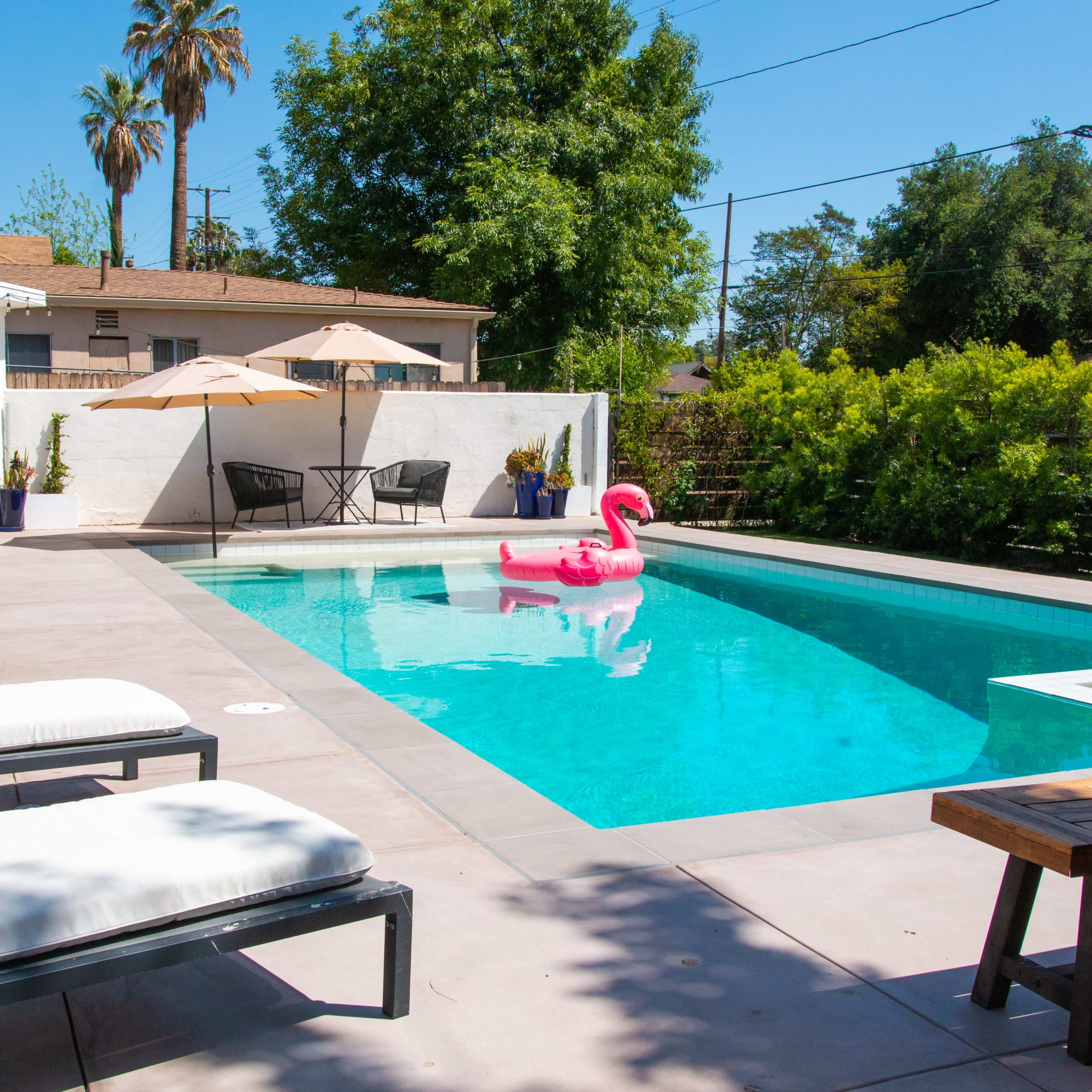 A clear swimming pool is surrounded by lounge chairs and a dining table, with a pink flamingo float in the water.