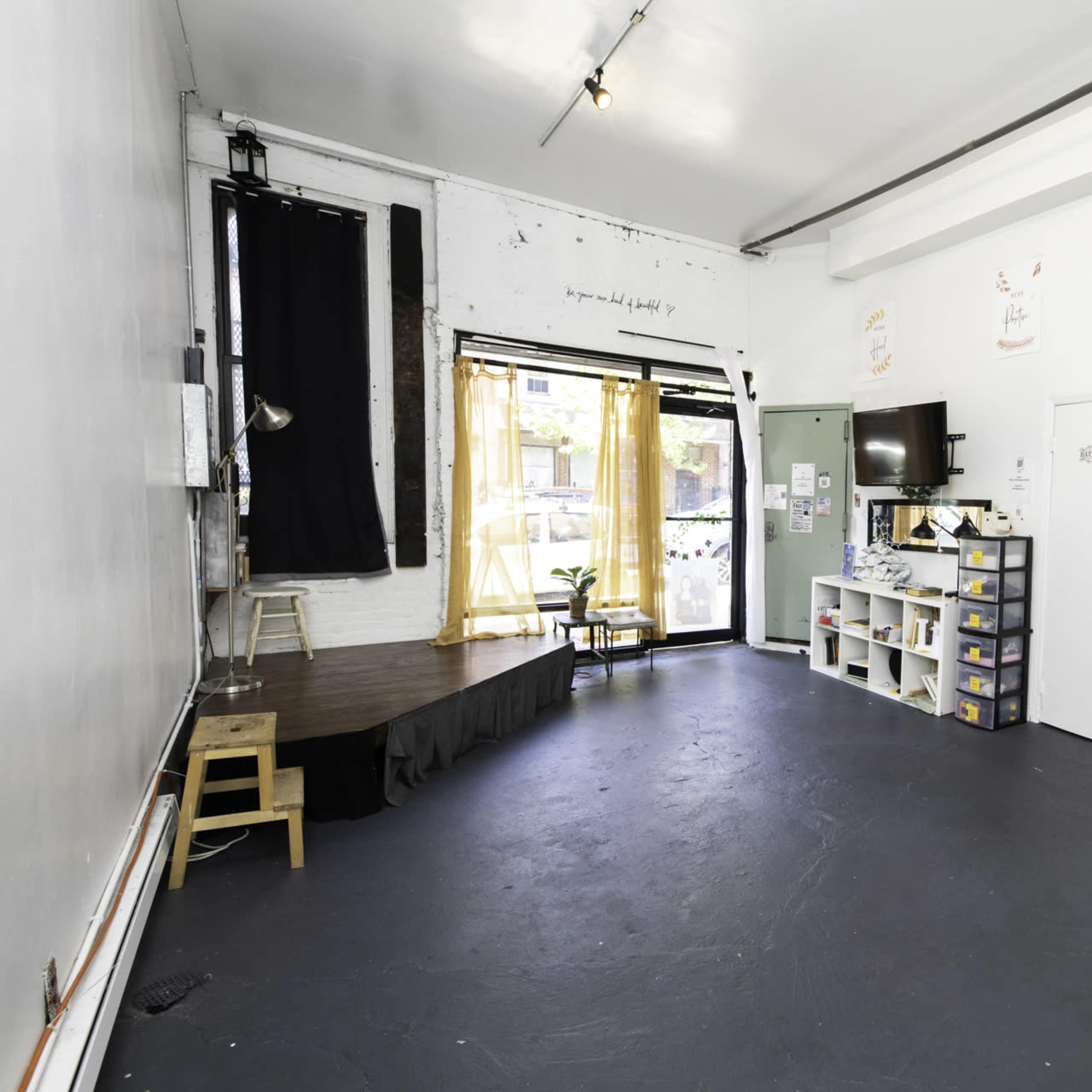 A spacious, empty room with a black stage area, a television on a wall, and storage boxes along one side.
