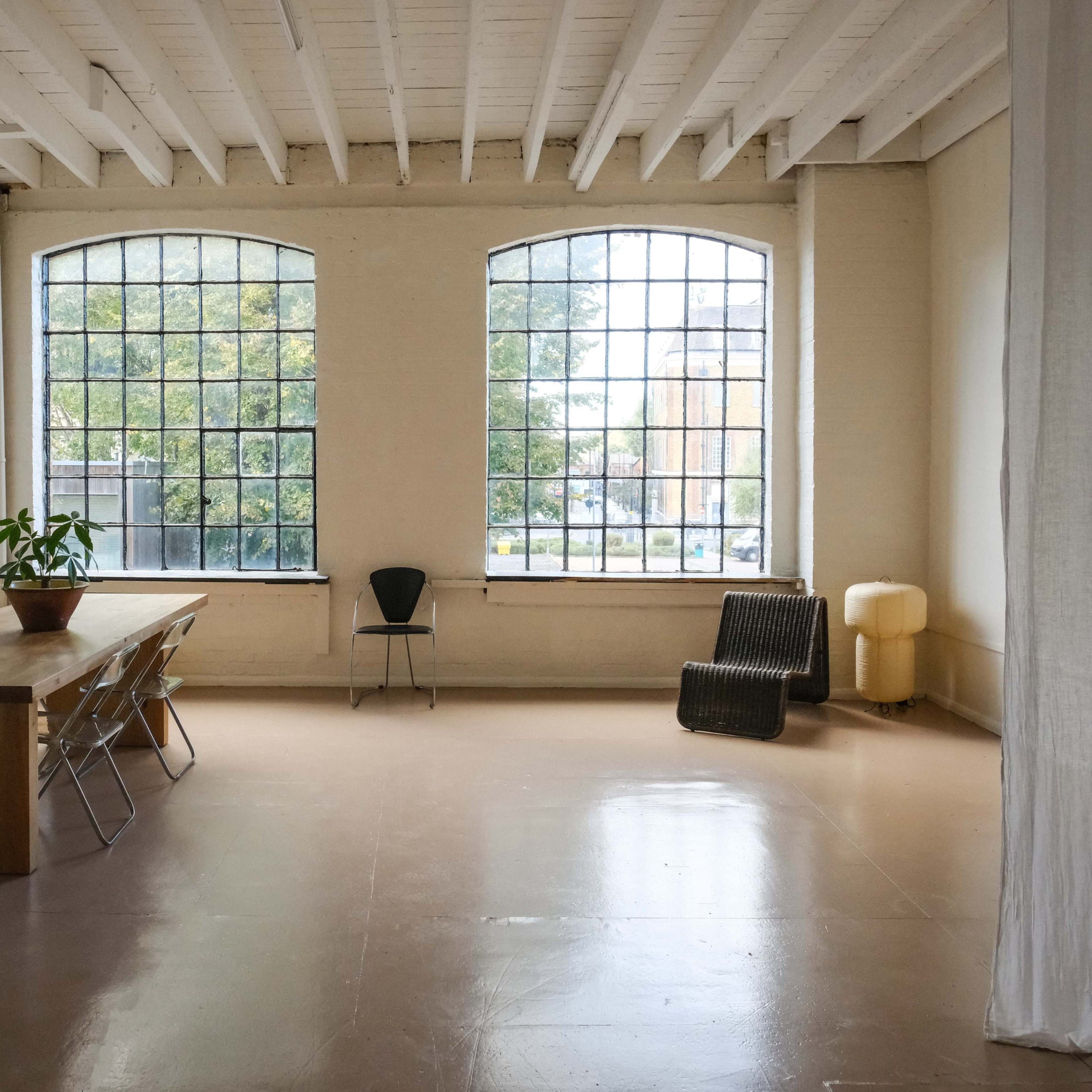 An empty room features a long wooden table, a chair, and large windows that allow natural light to enter.