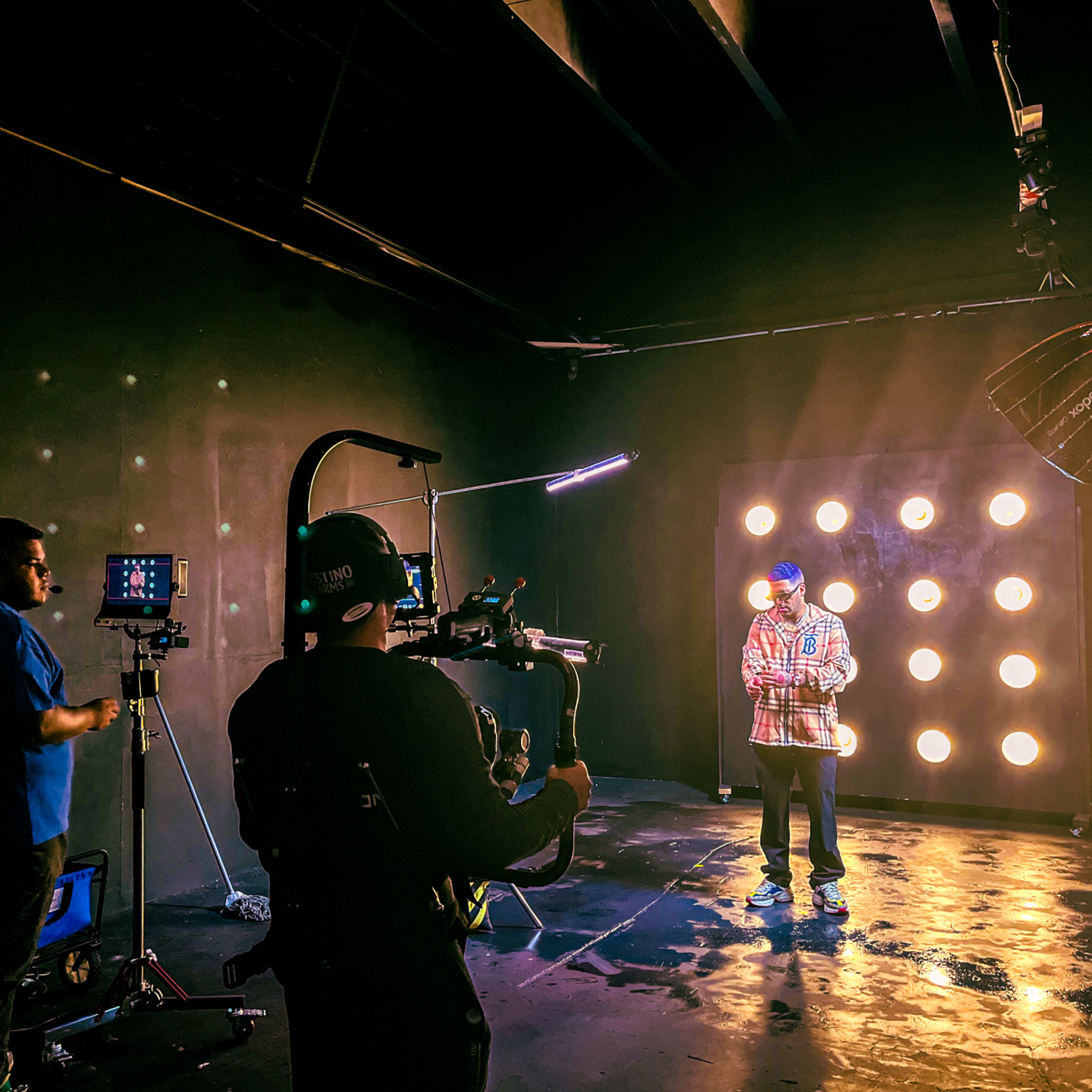 A person stands in a studio setting with a light fixture behind them, while two crew members operate filming equipment.