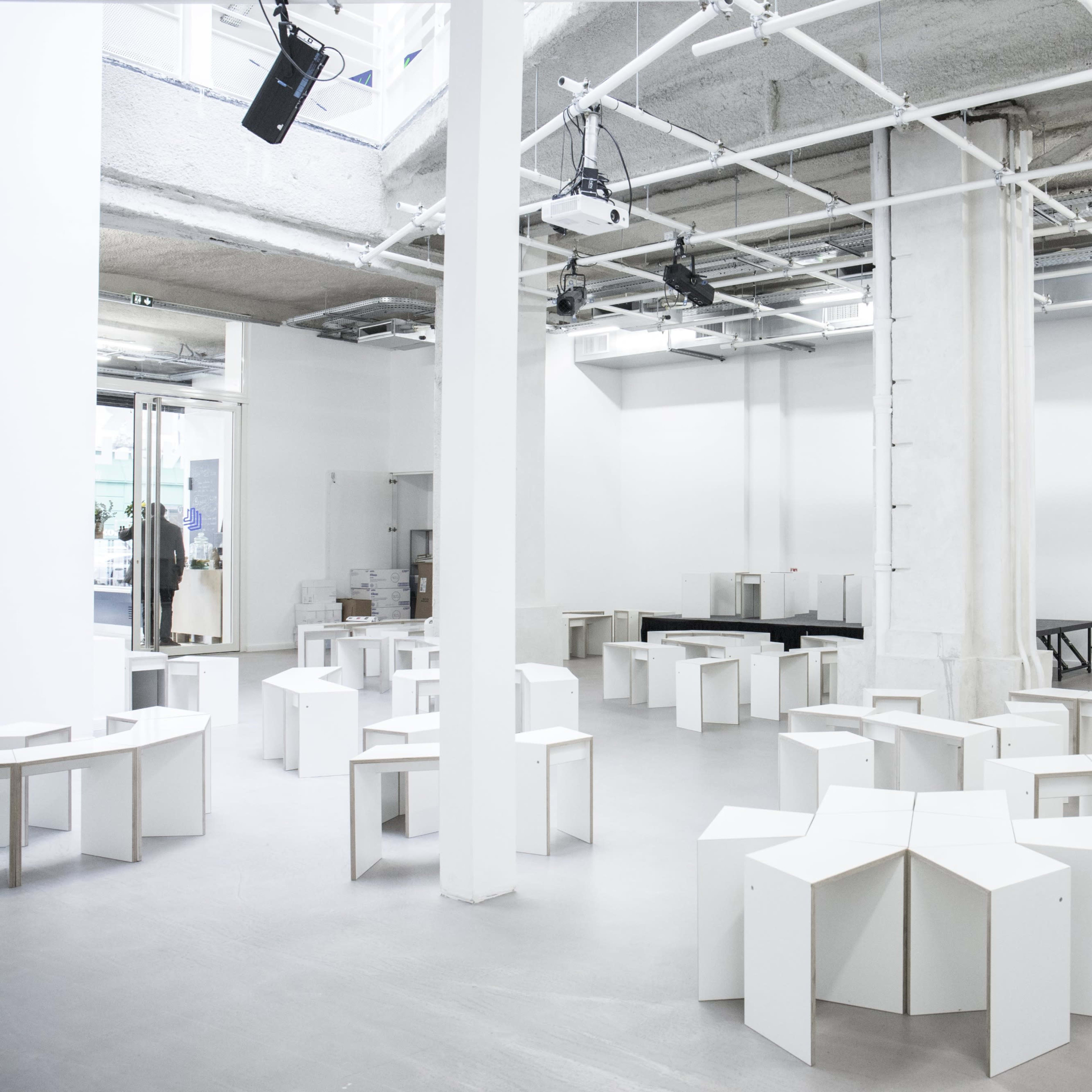 A spacious, empty interior with white walls and multiple angular, white tables arranged in a circular pattern.