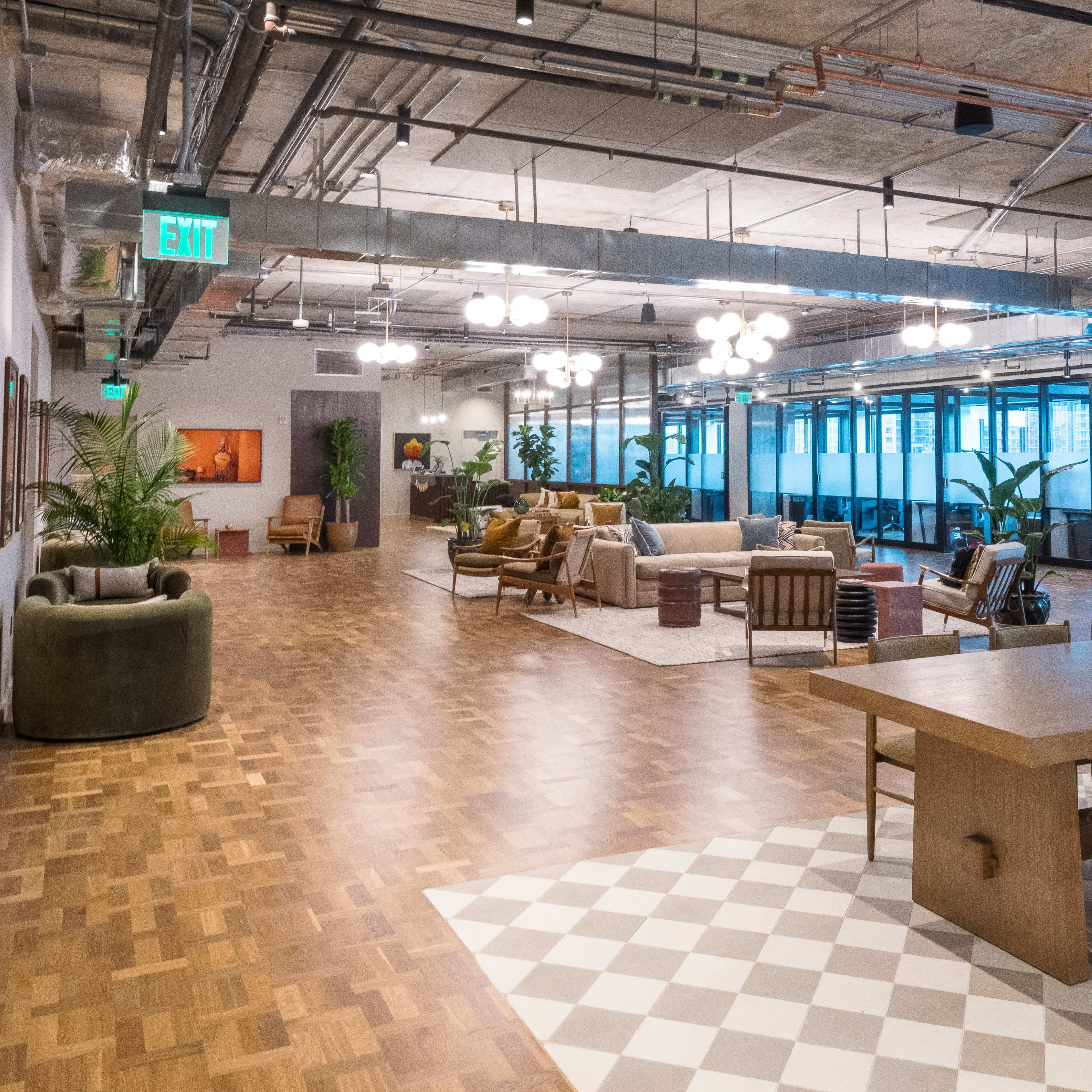 A modern office interior with various seating areas, a dining table, and large windows, featuring wooden floors and contemporary lighting.