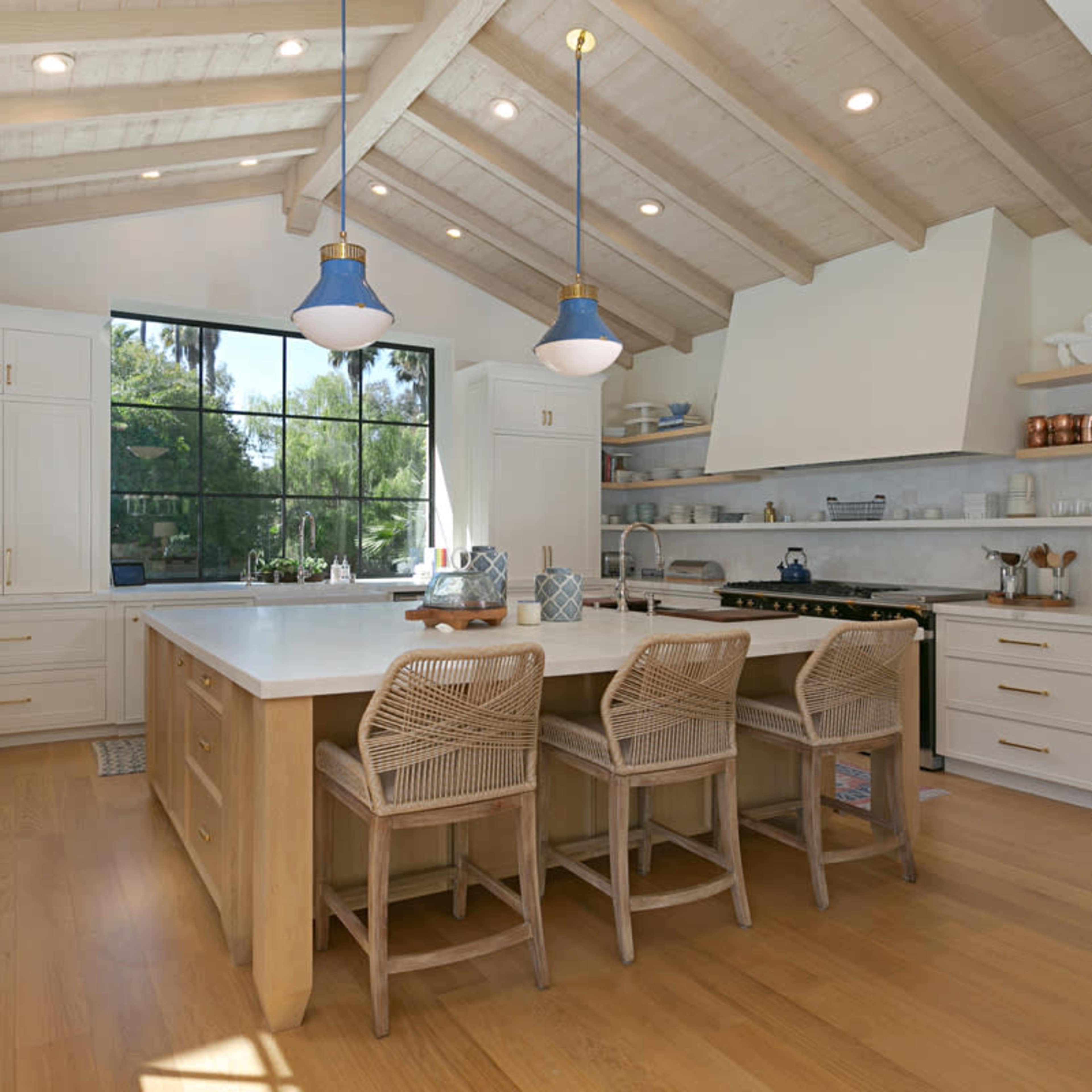 A spacious kitchen features a large island with three stools, white cabinetry, and wooden beams overhead, illuminated by pendant lights.
