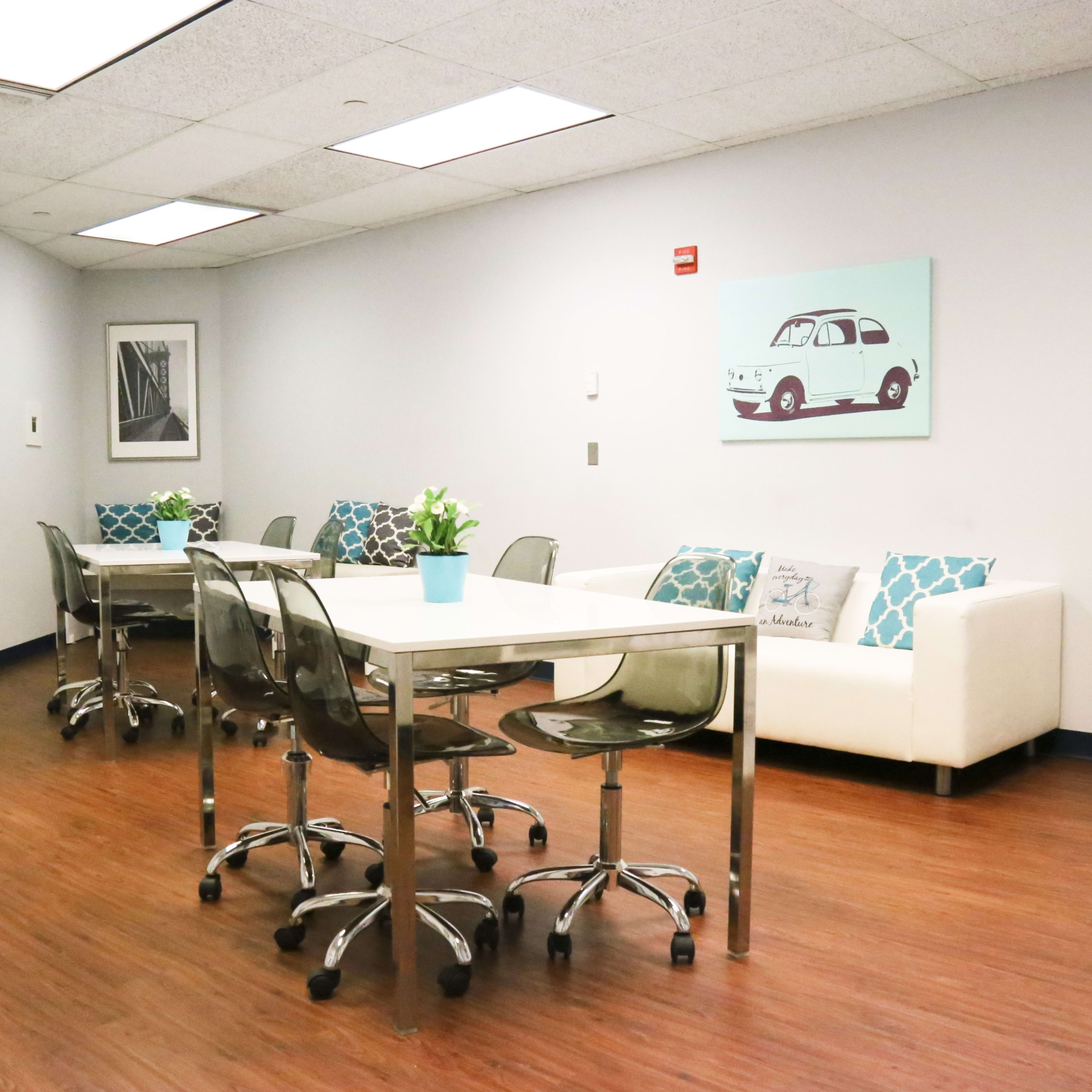 A modern office space featuring two glass tables surrounded by chairs, a white couch with decorative pillows, and wall art.