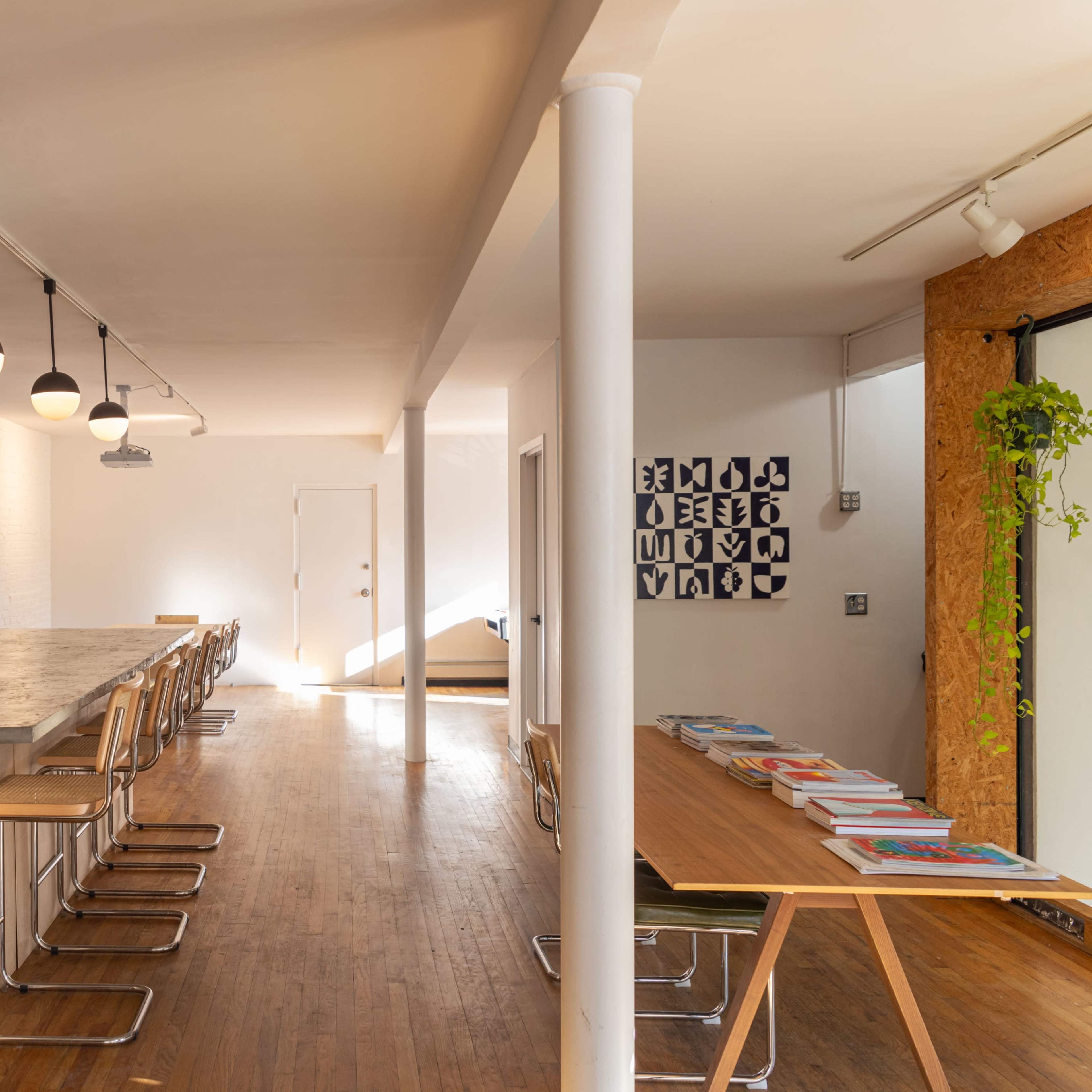 A spacious interior with a long wooden table and bar stools on one side, exposed brick walls, and a light-filled area featuring a modern design.