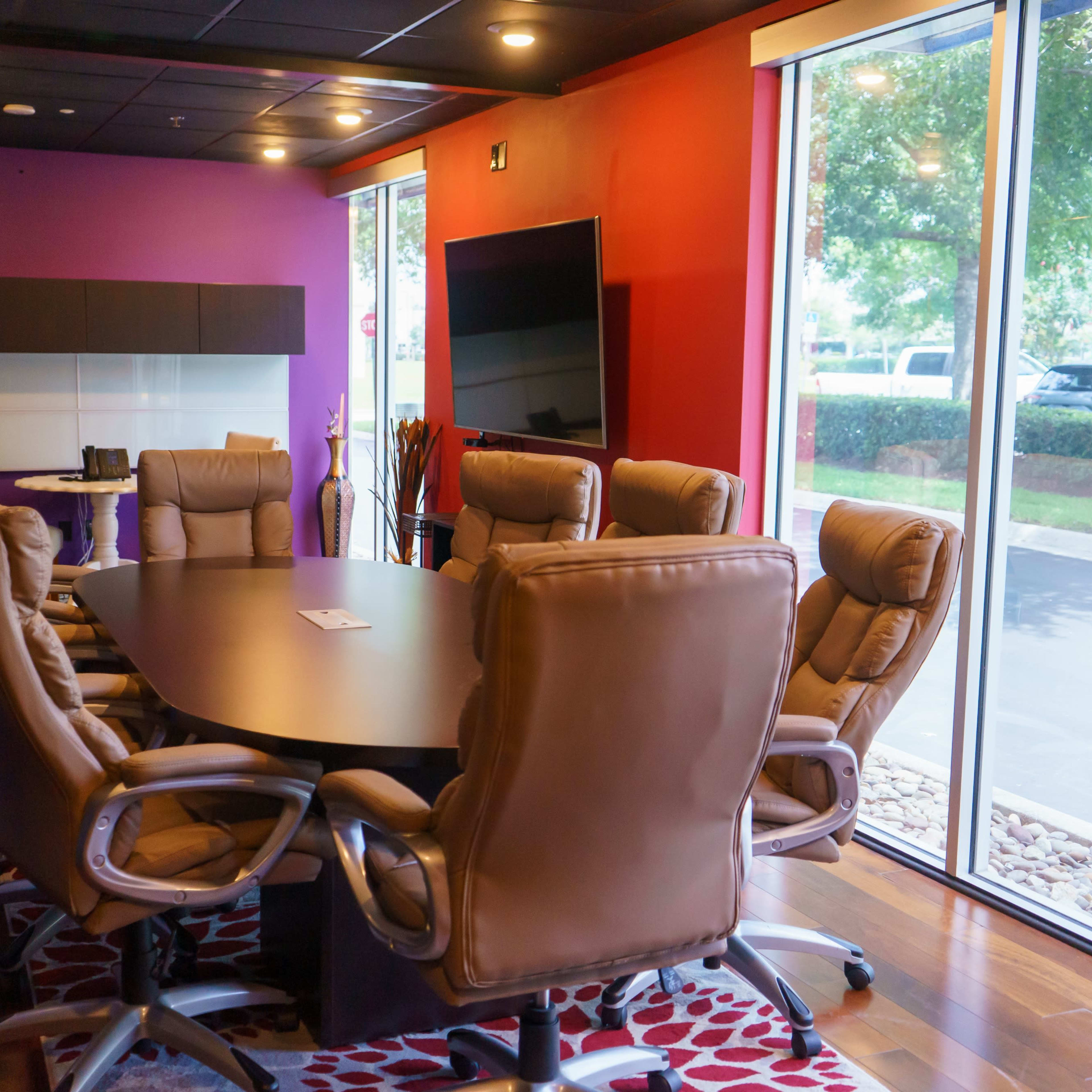 A modern conference room features a large oval table surrounded by ergonomic chairs, with a purple and red accent wall and large windows overlooking a landscaped area.