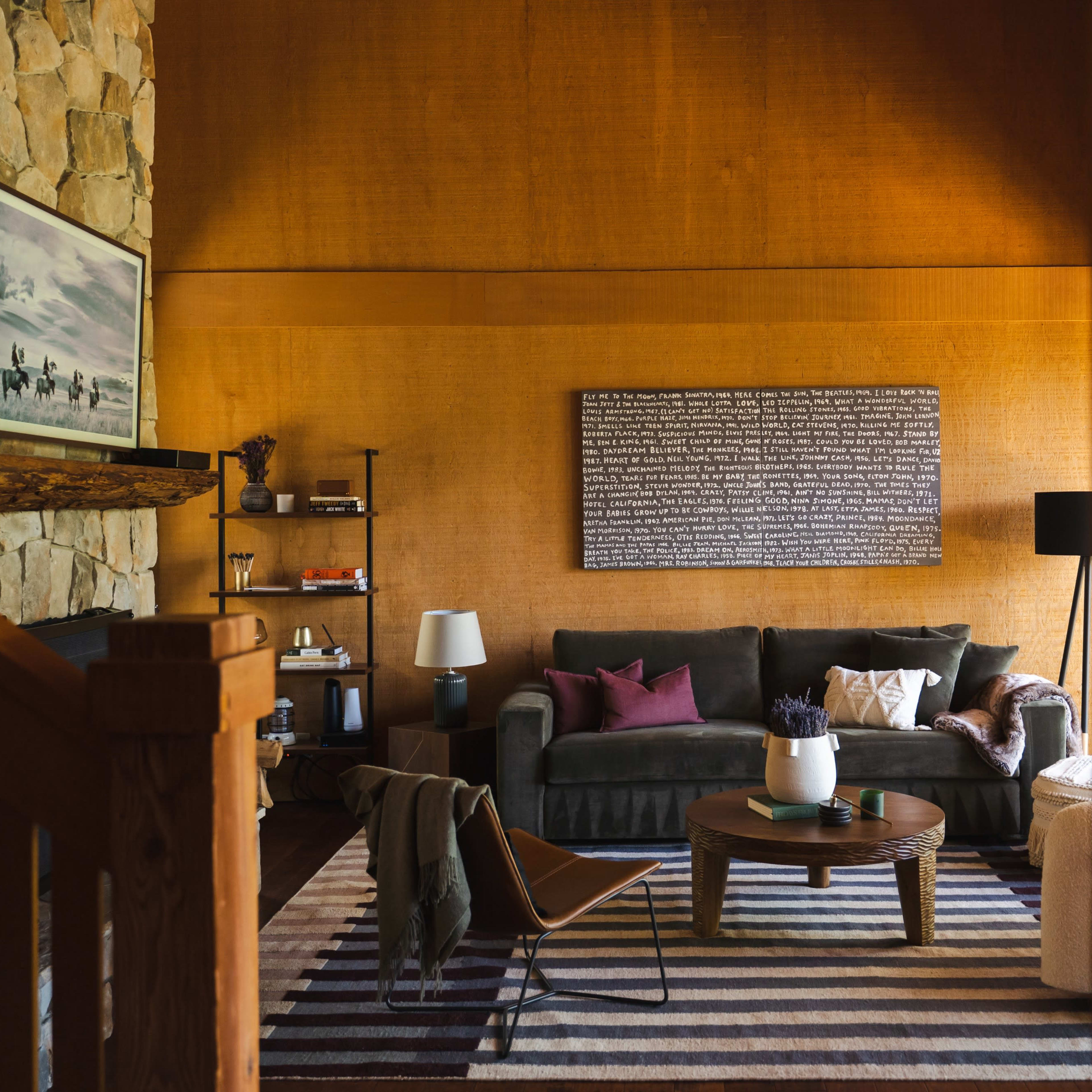 A cozy living room with a stone fireplace, a dark sofa, a round coffee table, and a large wall art piece, all accented by warm wood tones and a striped area rug.