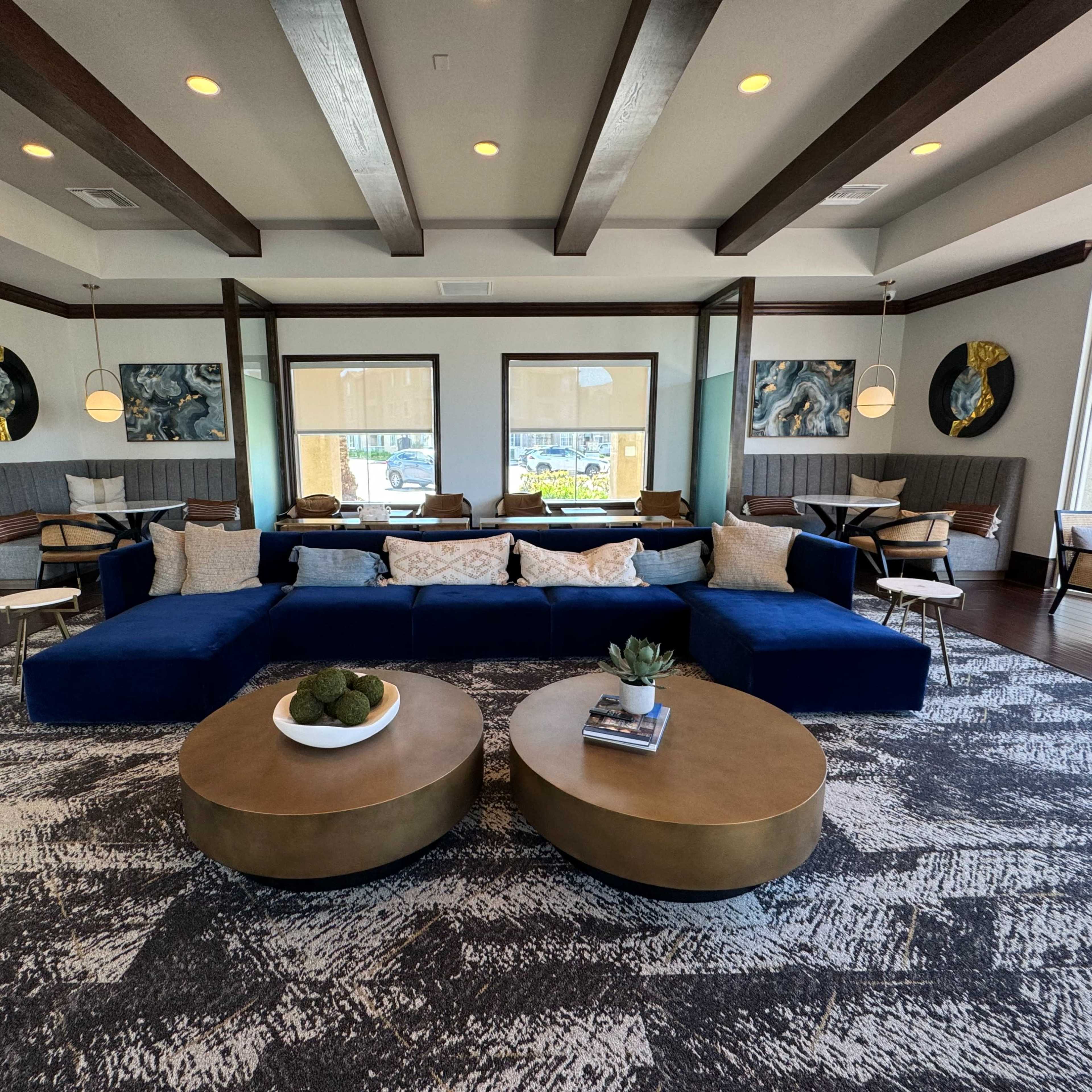 A modern lounge area with navy blue sectional sofas, round coffee tables, and large windows framed by decorative artwork and comfortable seating.