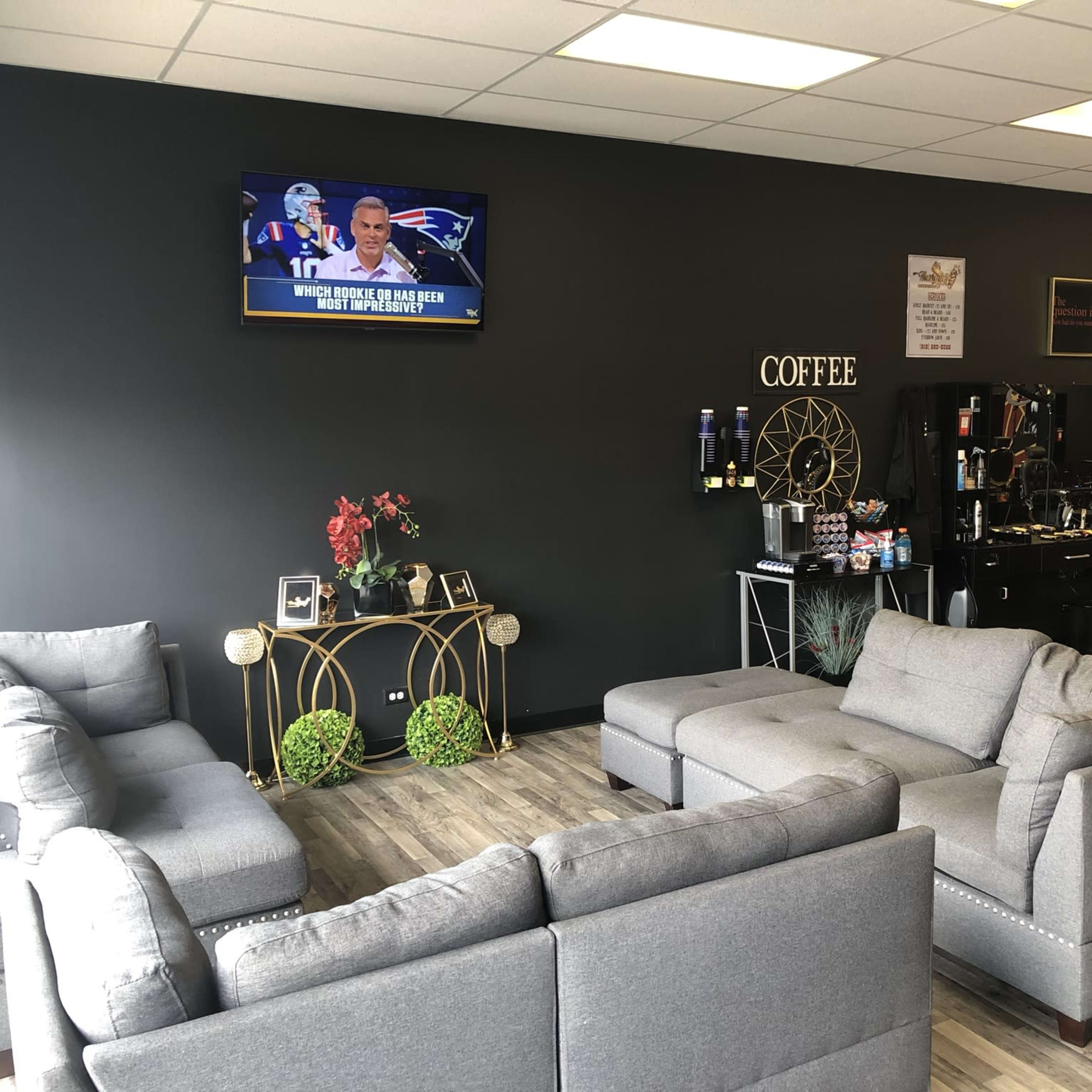 A waiting area with gray sofas, a wall-mounted television displaying a sports program, and a coffee station in a modern barber shop.