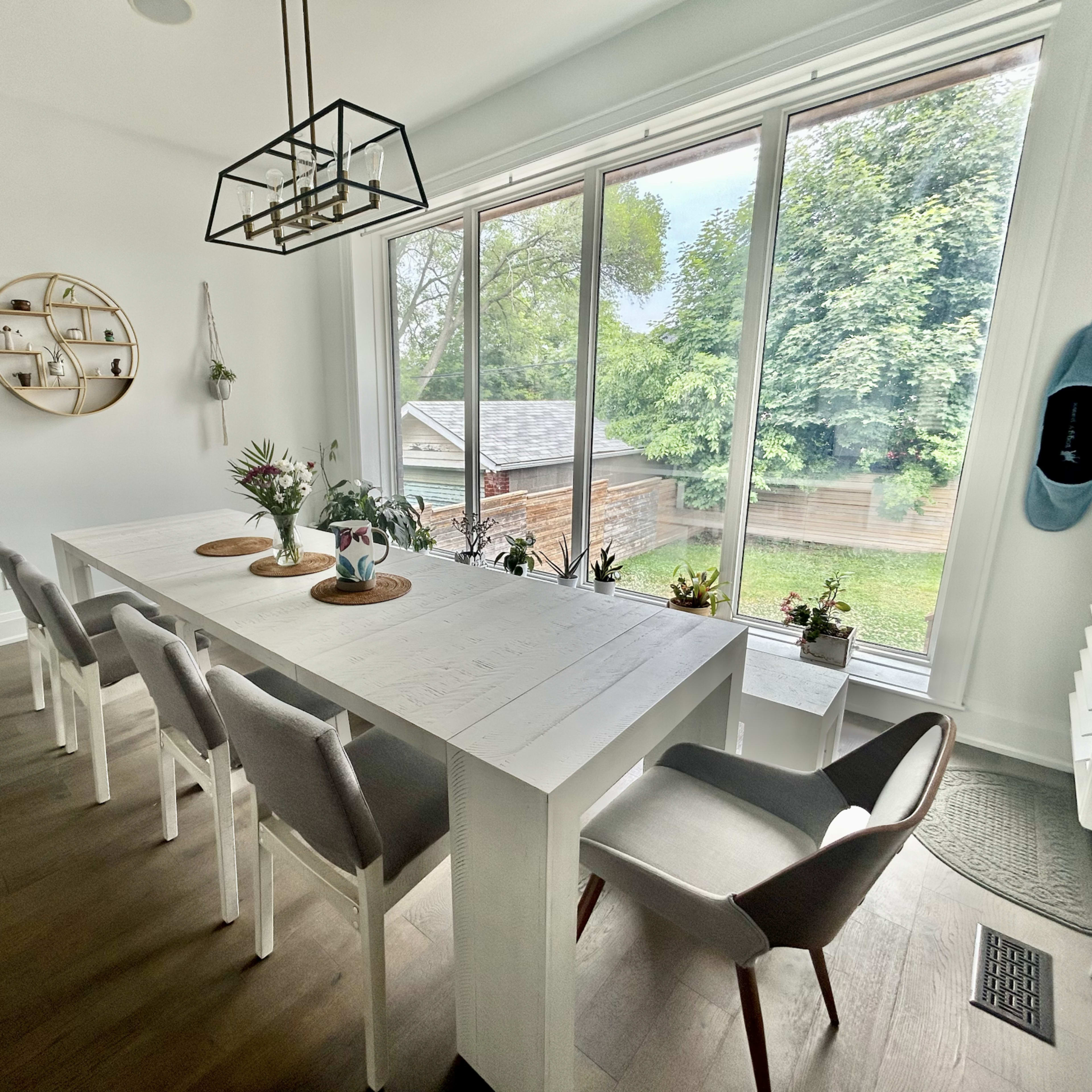A bright dining area features a long white table with gray chairs, surrounded by large windows offering a view of a green outdoor space.