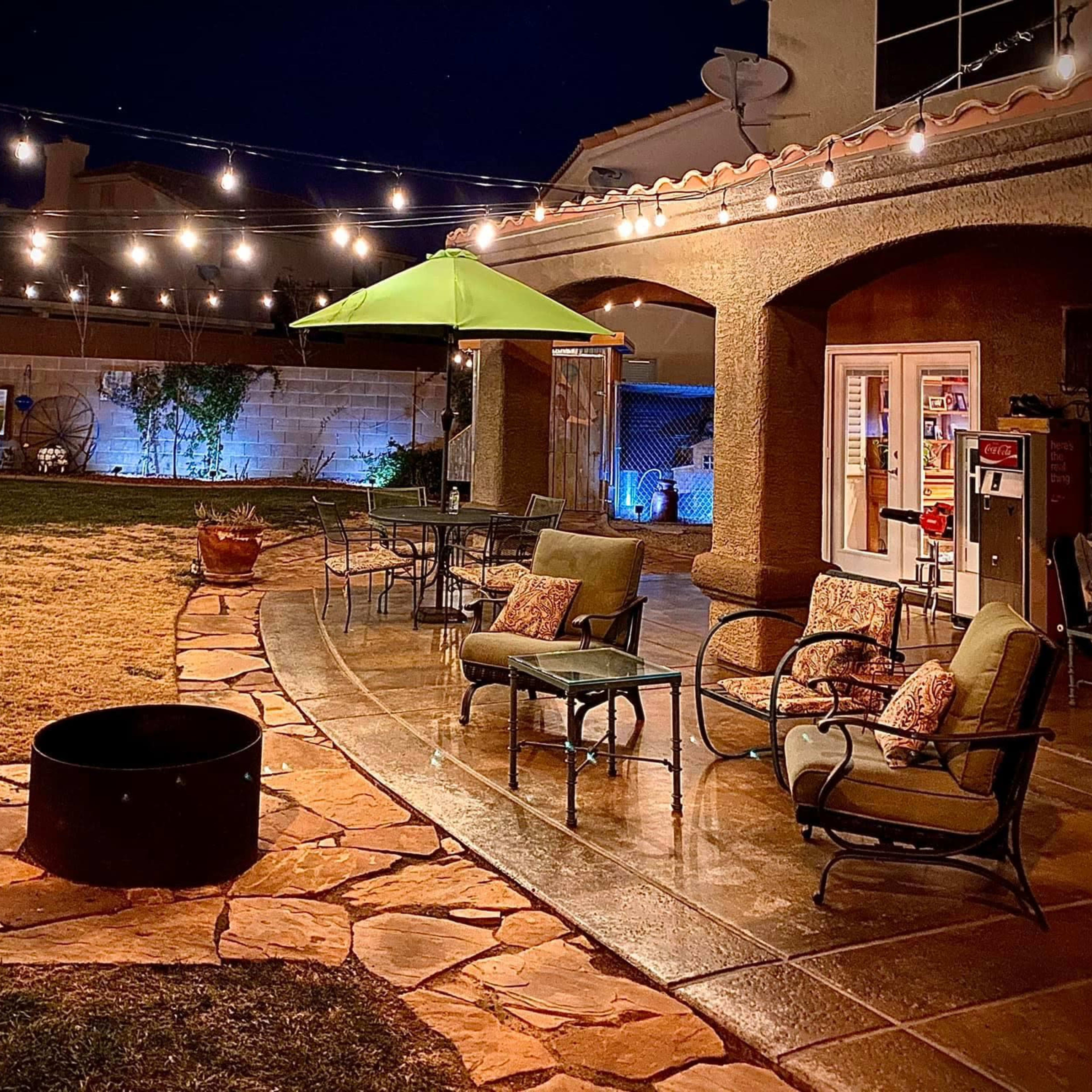 A backyard patio at night with string lights overhead, a green umbrella, seating arrangements, and a stone fire pit.