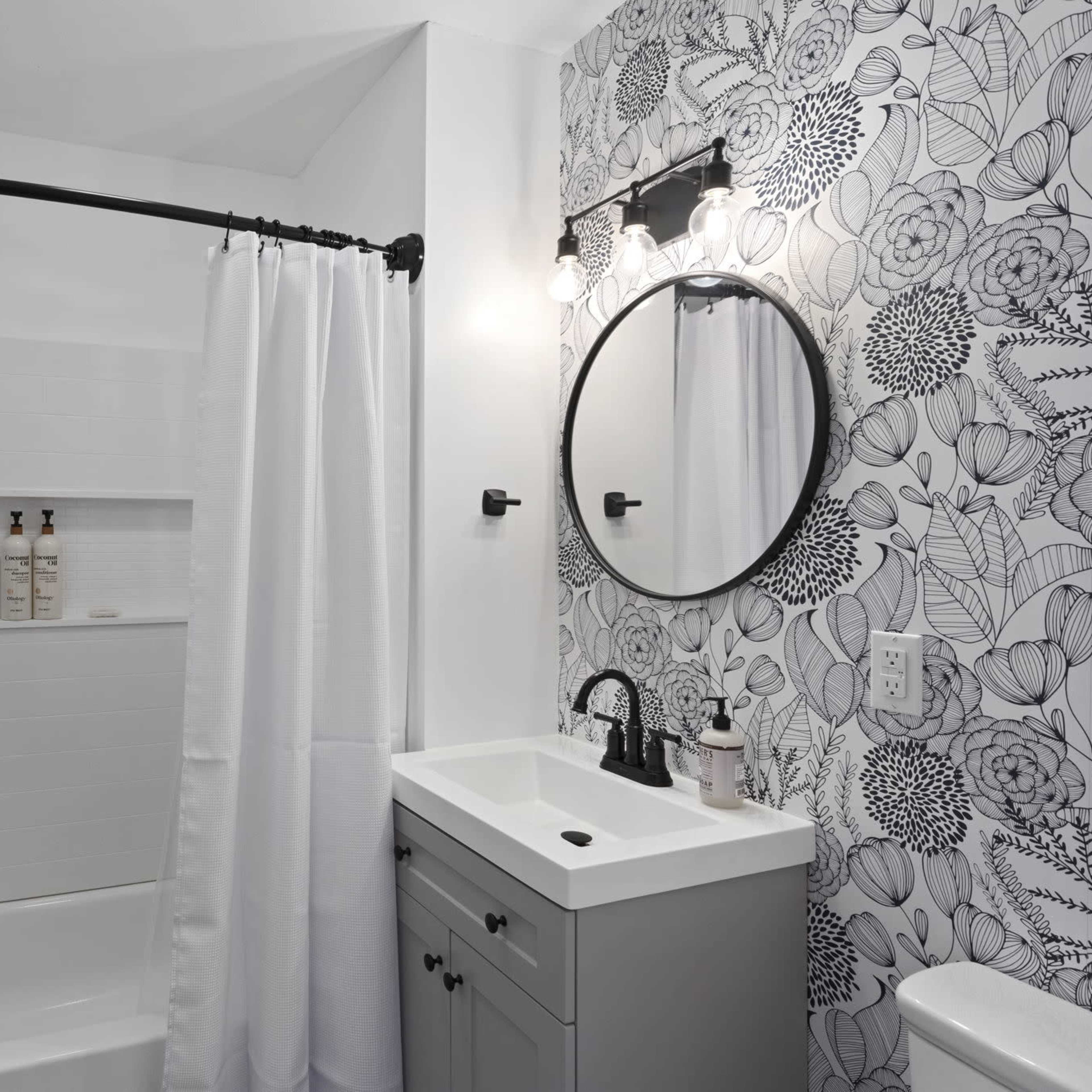 A modern bathroom featuring a shower with a white curtain, a round mirror above a gray vanity, and floral wallpaper.