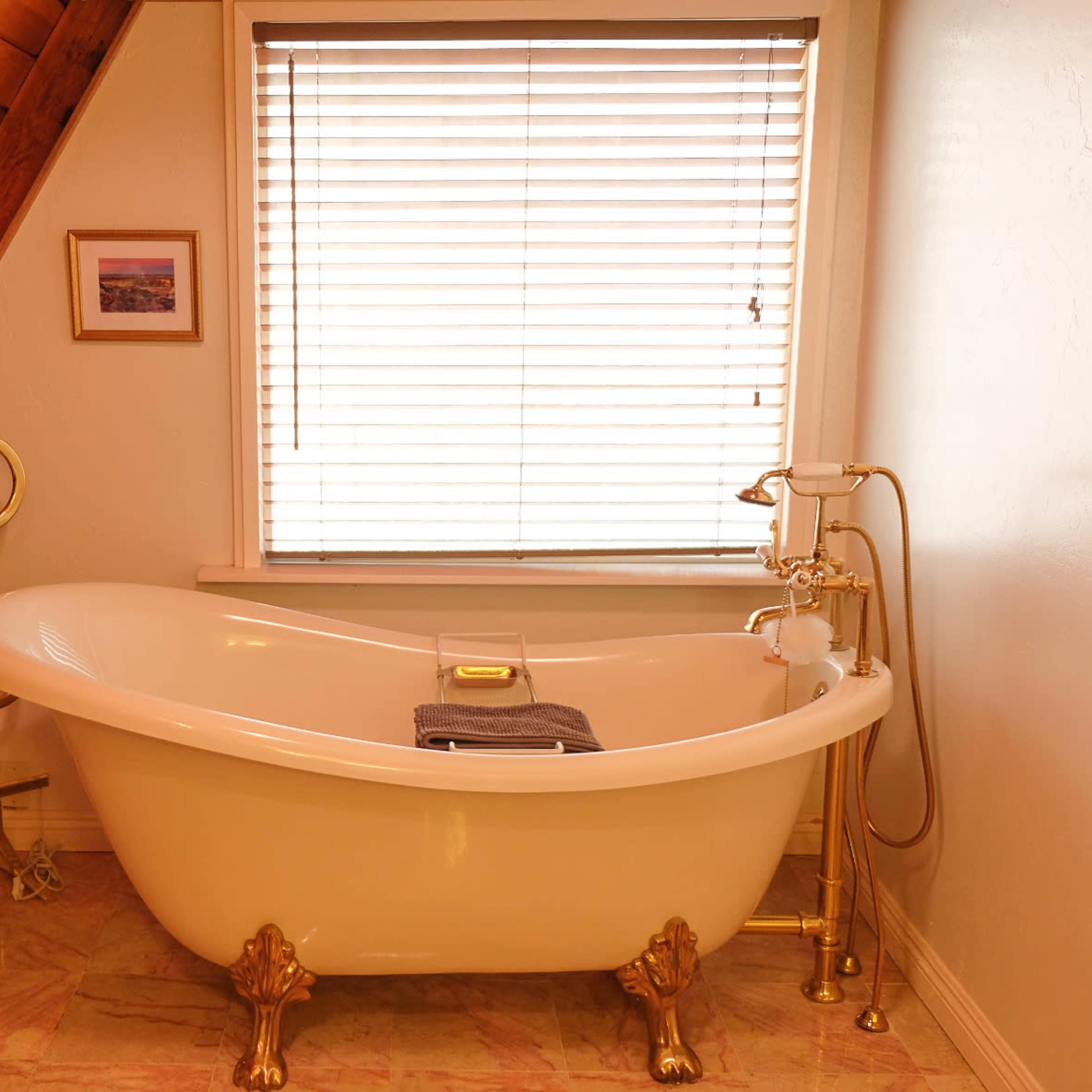 The scene shows a vintage-style bathtub with gold feet, positioned next to a window with blinds, in a room featuring wooden beams and light-colored walls.