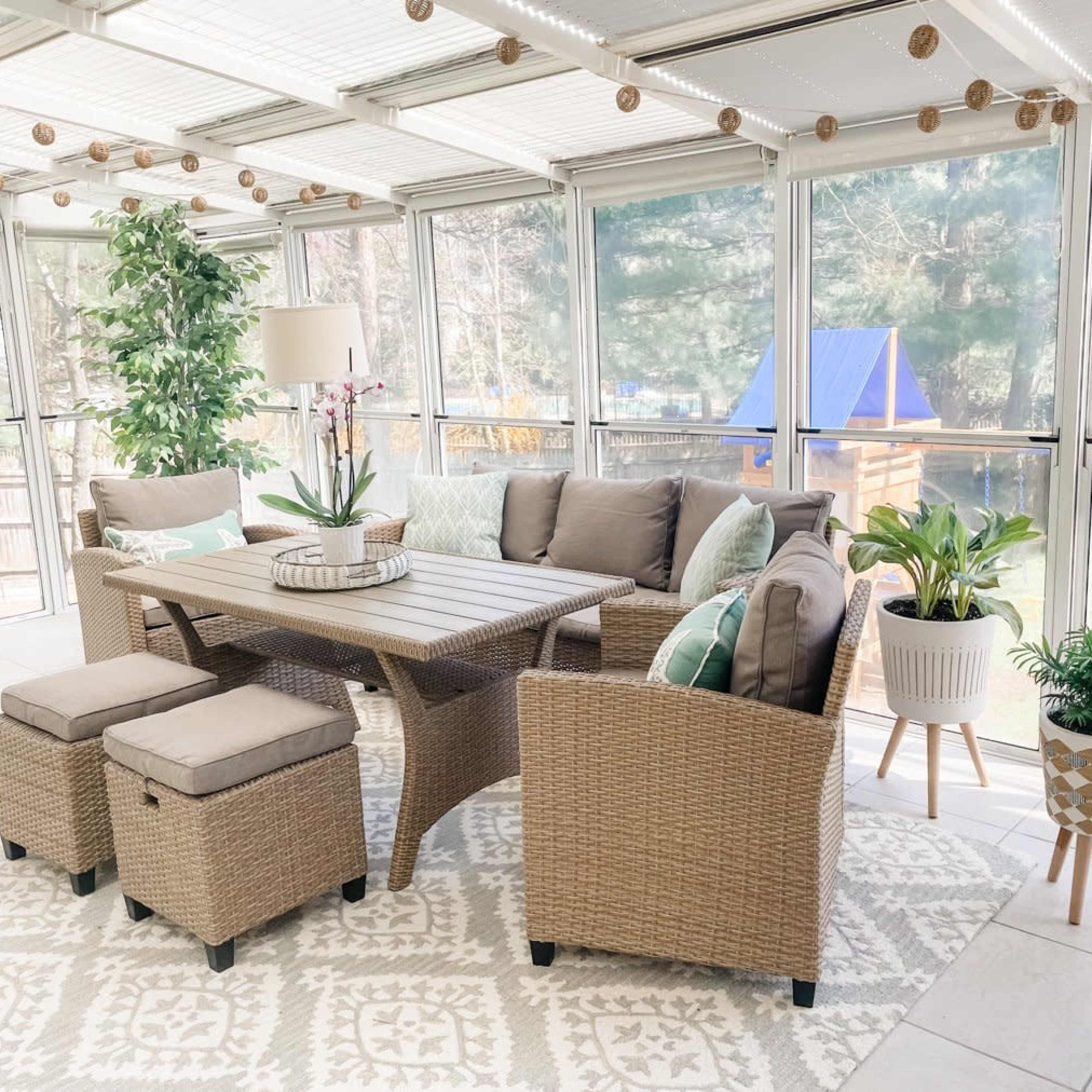 A sunroom features a wicker table and seating with decorative cushions, surrounded by greenery and bright natural light.