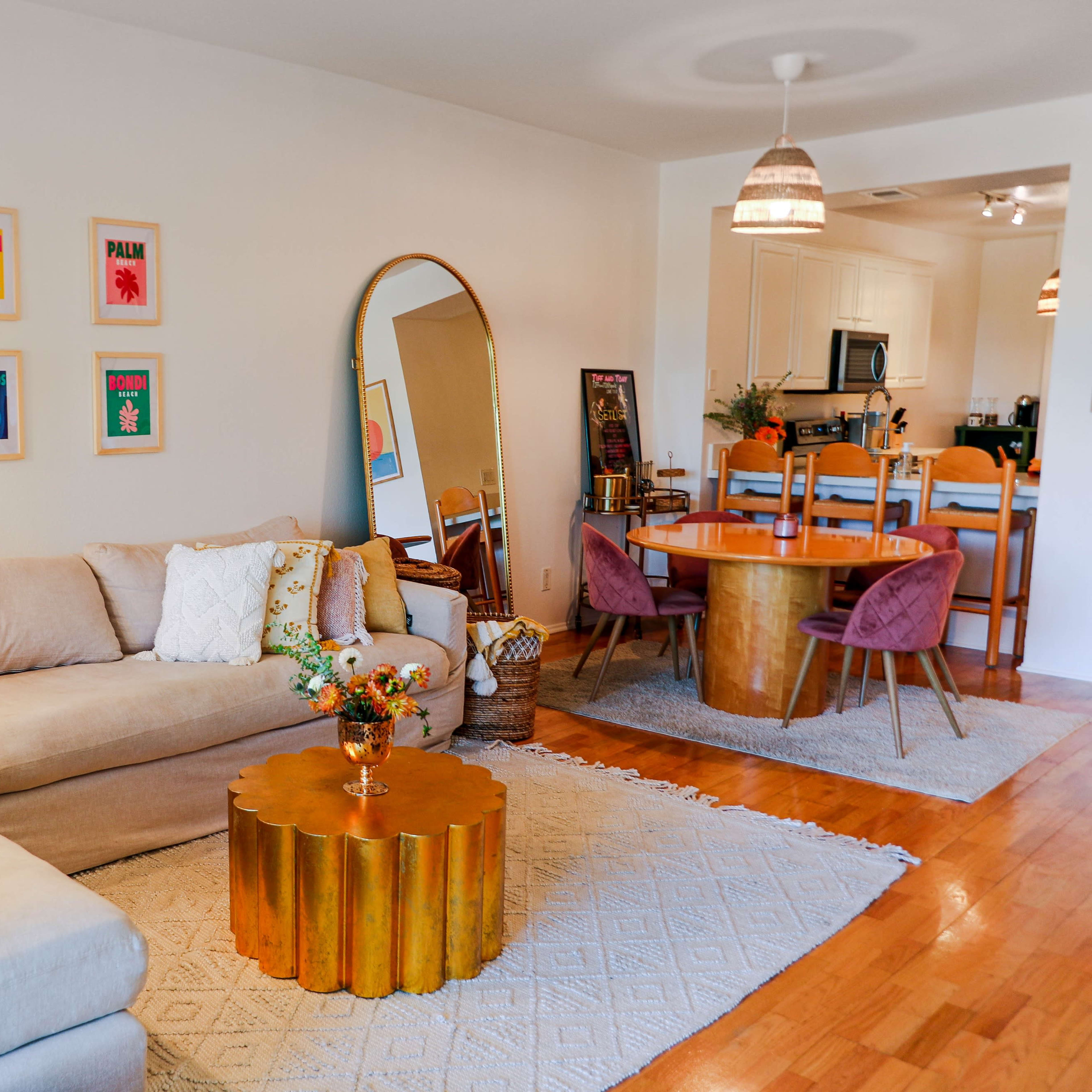 A bright living room with a beige sectional sofa, a round gold coffee table, colorful framed artwork on the wall, and a dining area with a round table and chairs.