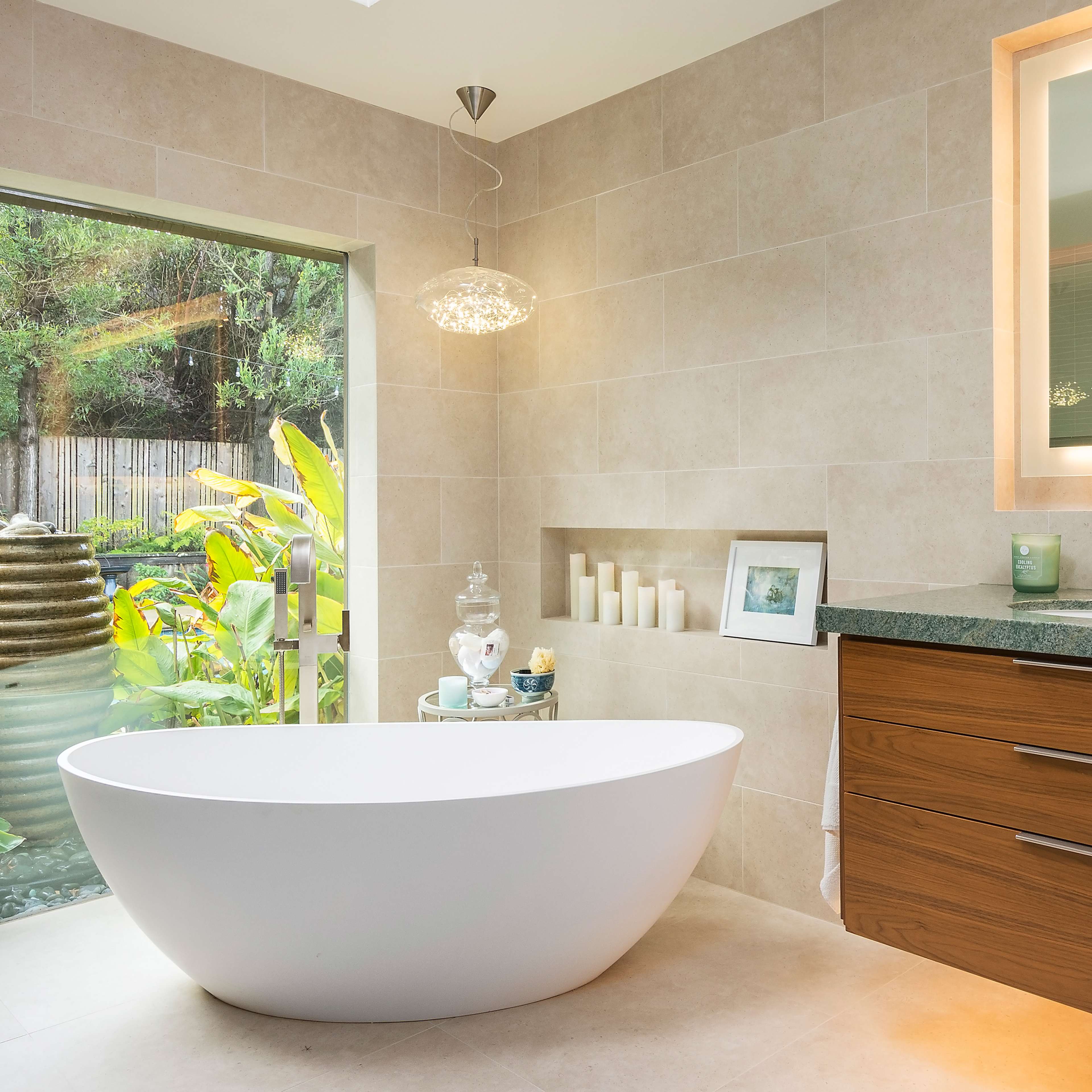 A modern bathroom features a freestanding white bathtub, wooden cabinetry, and large windows that provide a view of an outdoor garden.