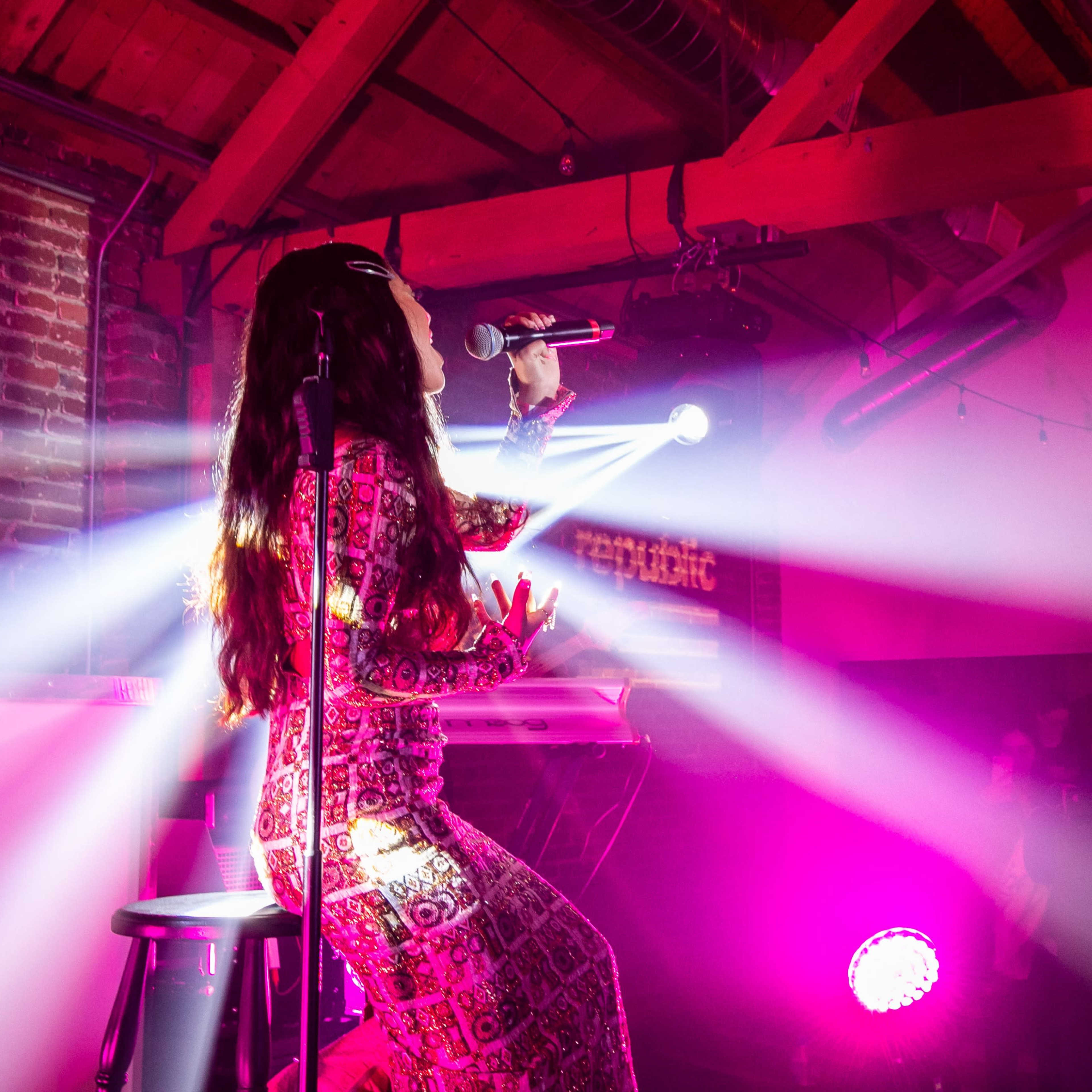 A singer performs on stage in a dimly lit venue with colorful lights and a crowd in the background.