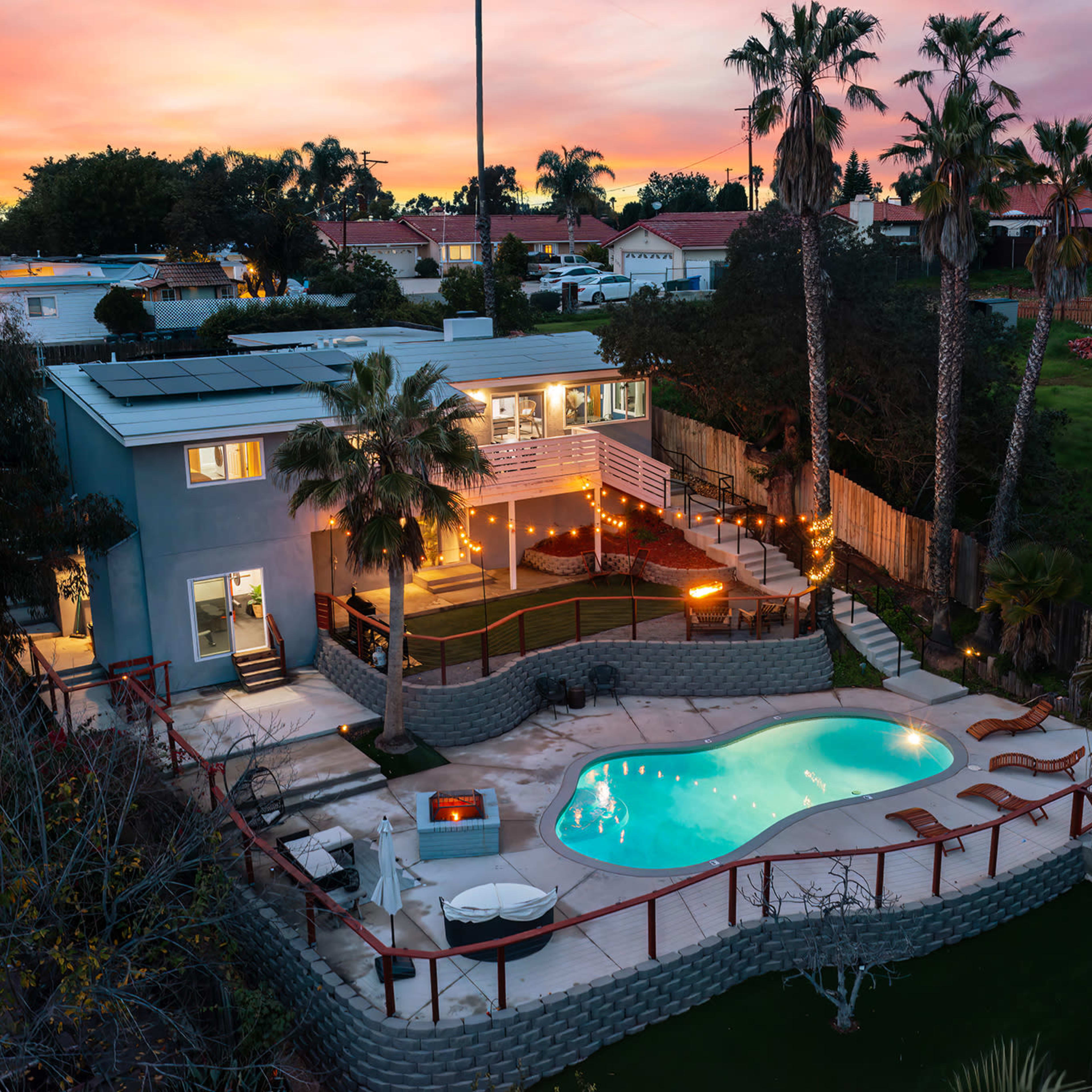 A modern house with a pool surrounded by palm trees, illuminated by lights at dusk.
