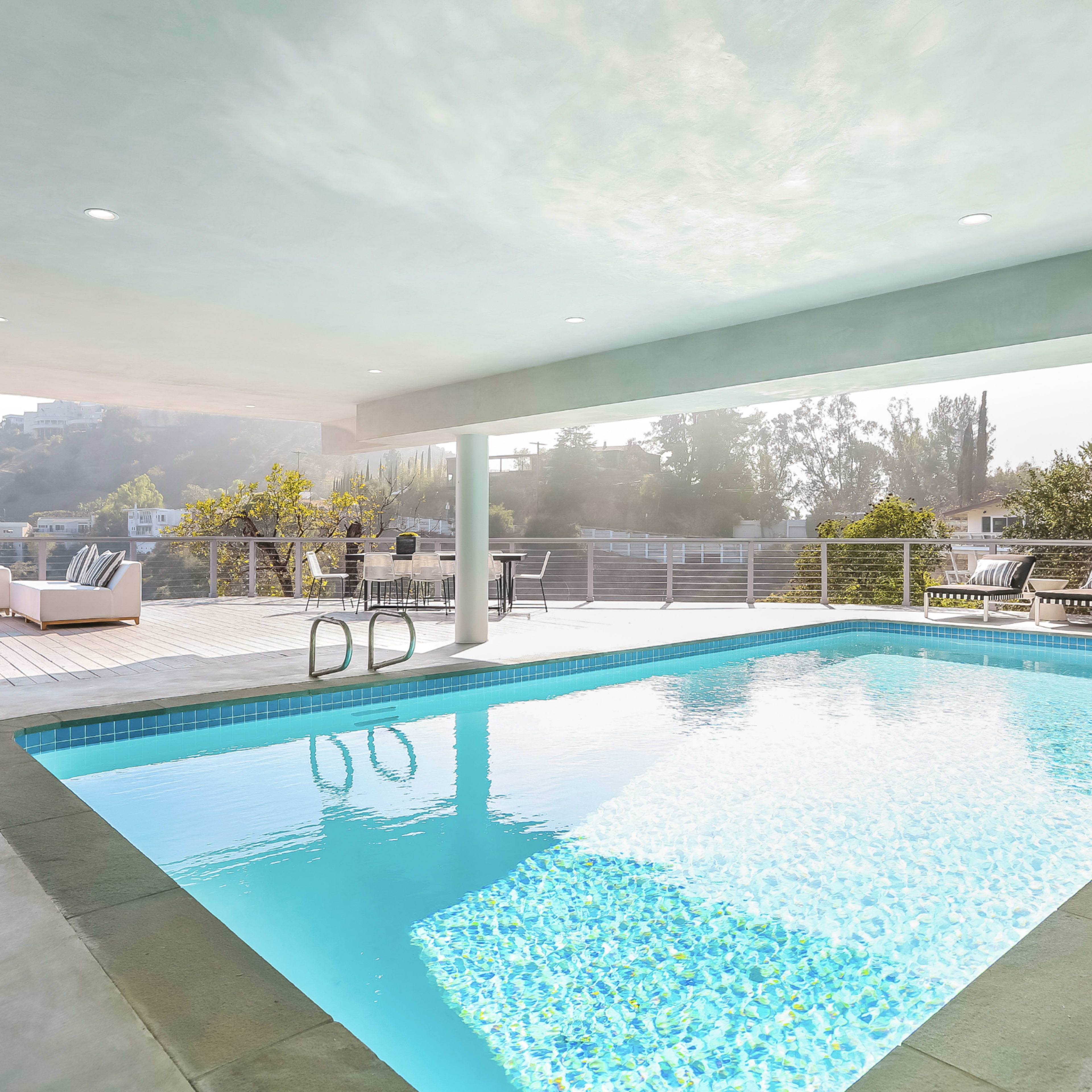 A modern outdoor area features a rectangular swimming pool surrounded by loungers and seating under a covered patio.