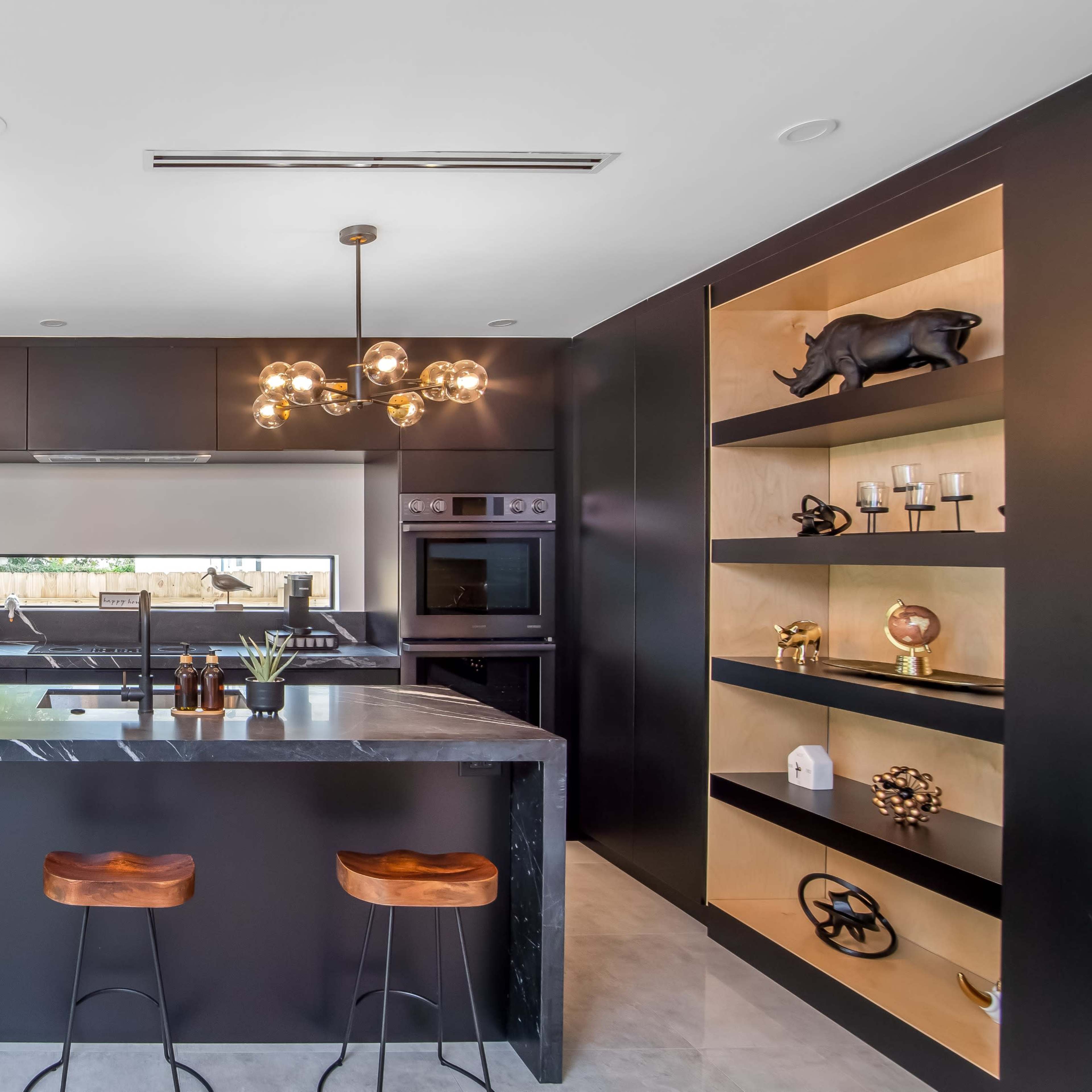 A modern kitchen featuring a large island with three bar stools, black cabinetry, and decorative shelves with various objects.