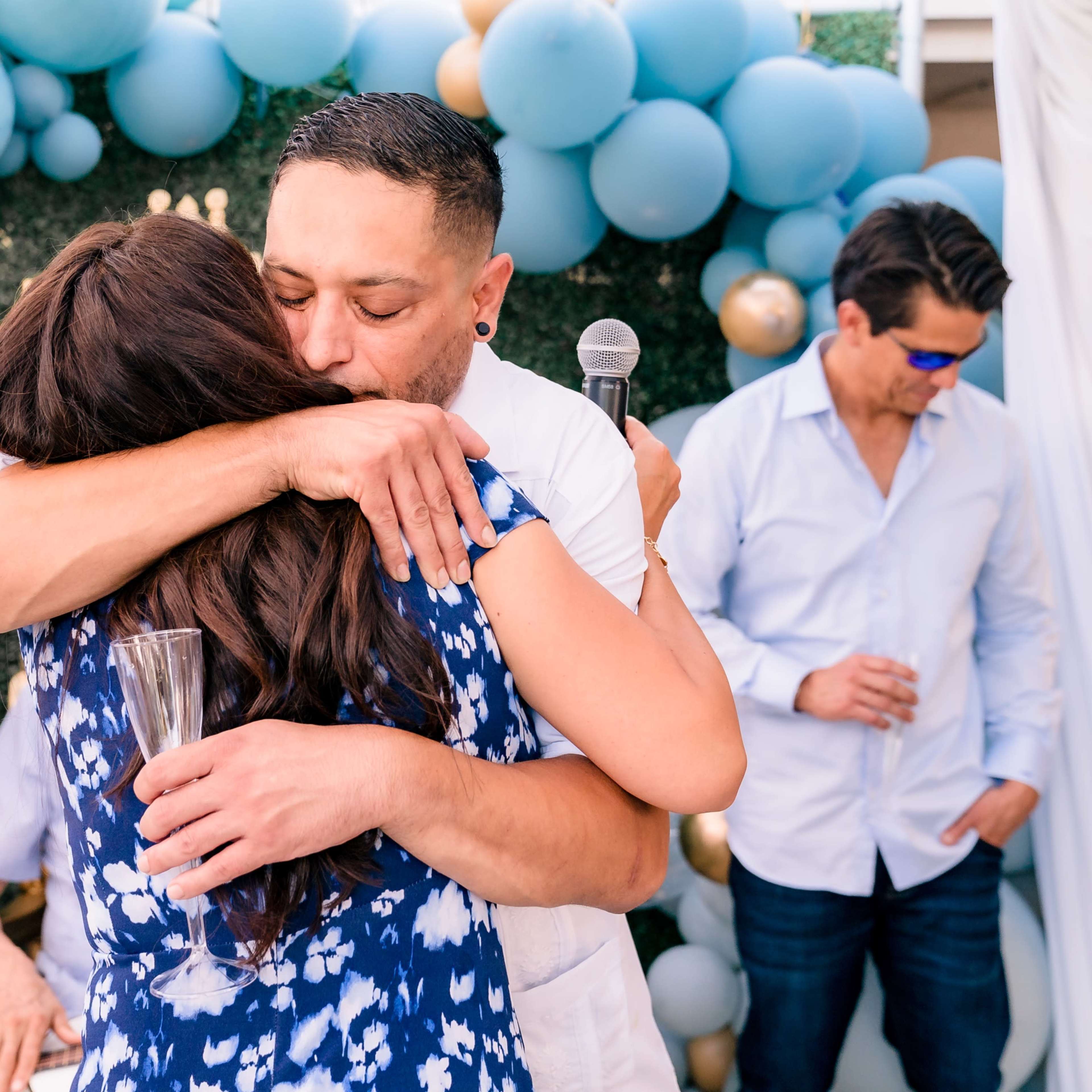 A man and woman embrace in celebration, surrounded by guests who are clapping and enjoying the event, with a backdrop of blue and gold balloons.